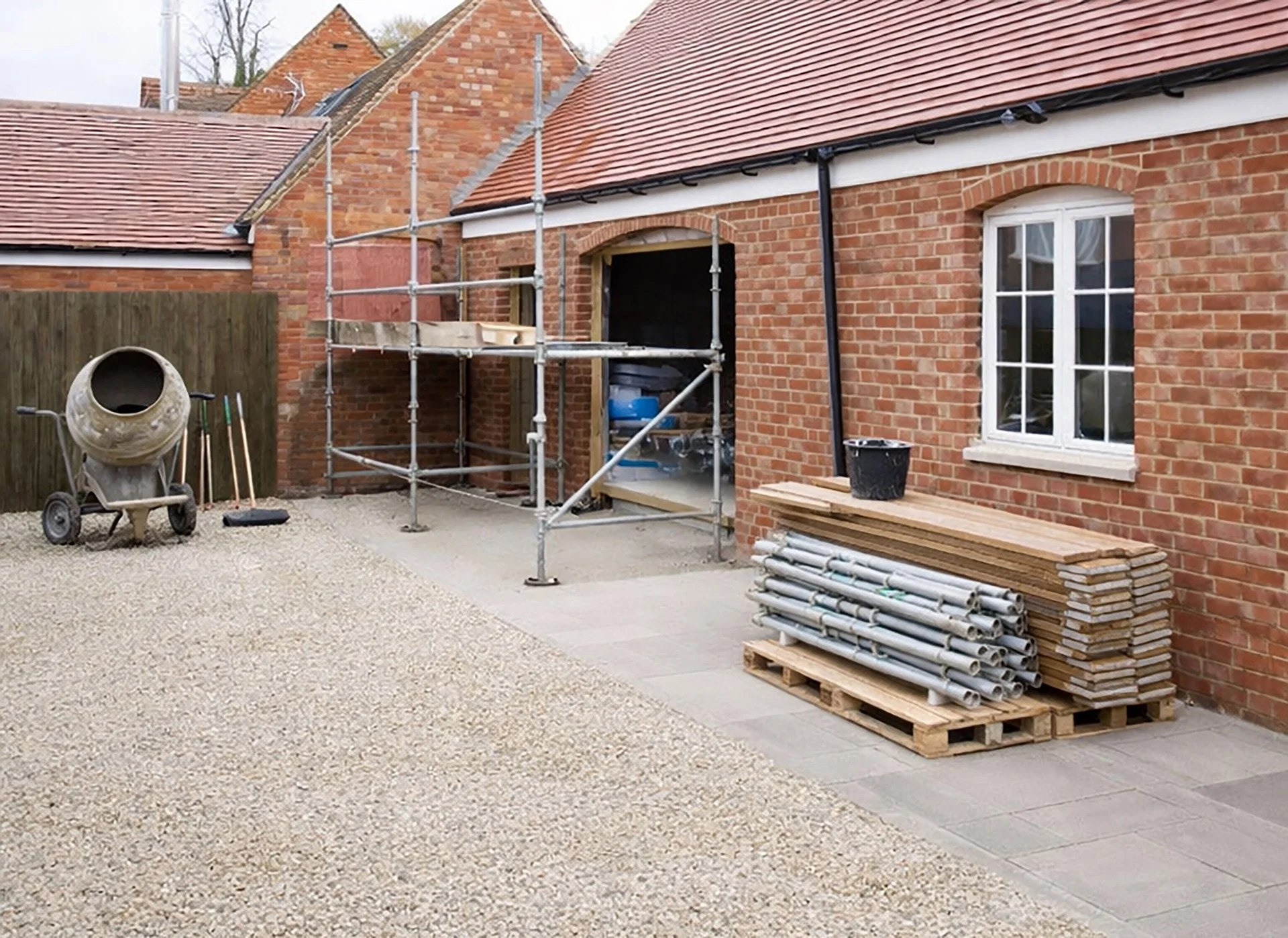 A red brick home undergoing renovation, shown with a cement mixer, scaffolding, and stacked building materials.