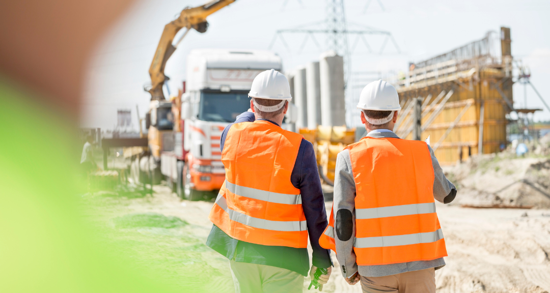 Two construction workers wearing white helmets and orange safety vests walking on a construction site with cranes and heavy machinery in the background.