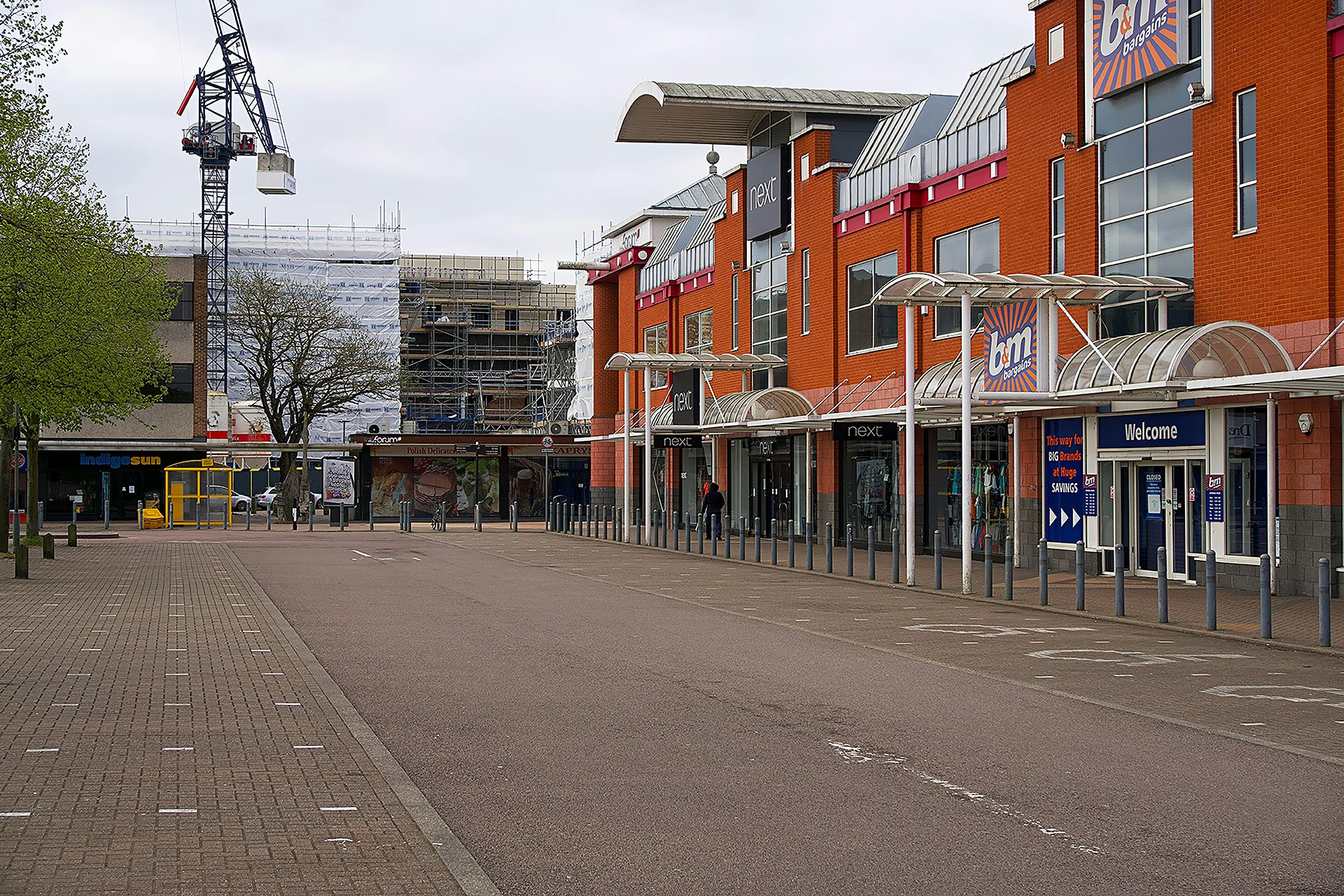 An outdoor wide shot of a retail park featuring a row of two-story brick storefronts on the right, including a "B&M" and a "Next."