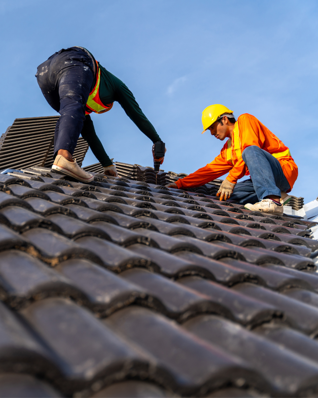 Two workers in safety gear installing or repairing roof tiles on a sloped roof under a clear blue sky.