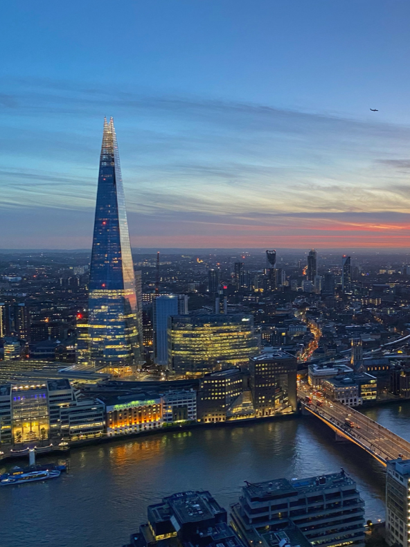 A city skyline at dusk featuring the Shard skyscraper, illuminated buildings, a river, and a bridge.
