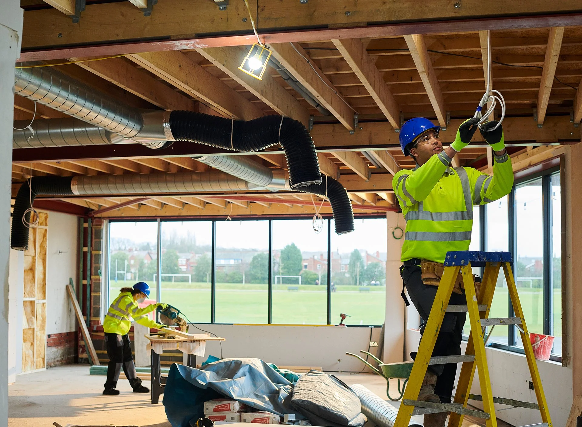 An electrician on a ladder installs wiring in a ceiling while another worker uses a circular saw in the background of a building under construction.