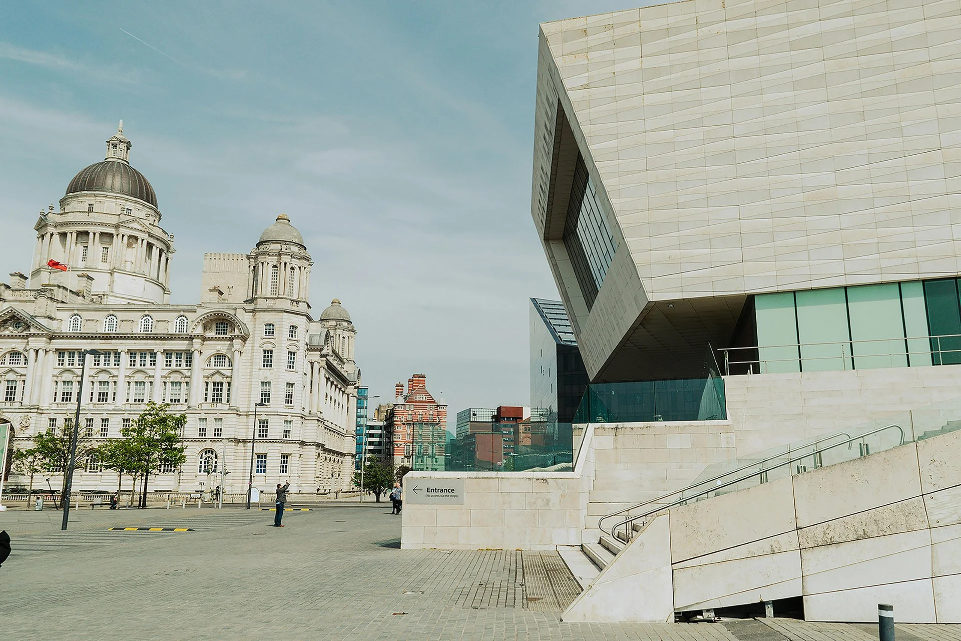 A modern local authority building with sharp angles stands opposite a domed historic landmark. The open square sits under a clear sky