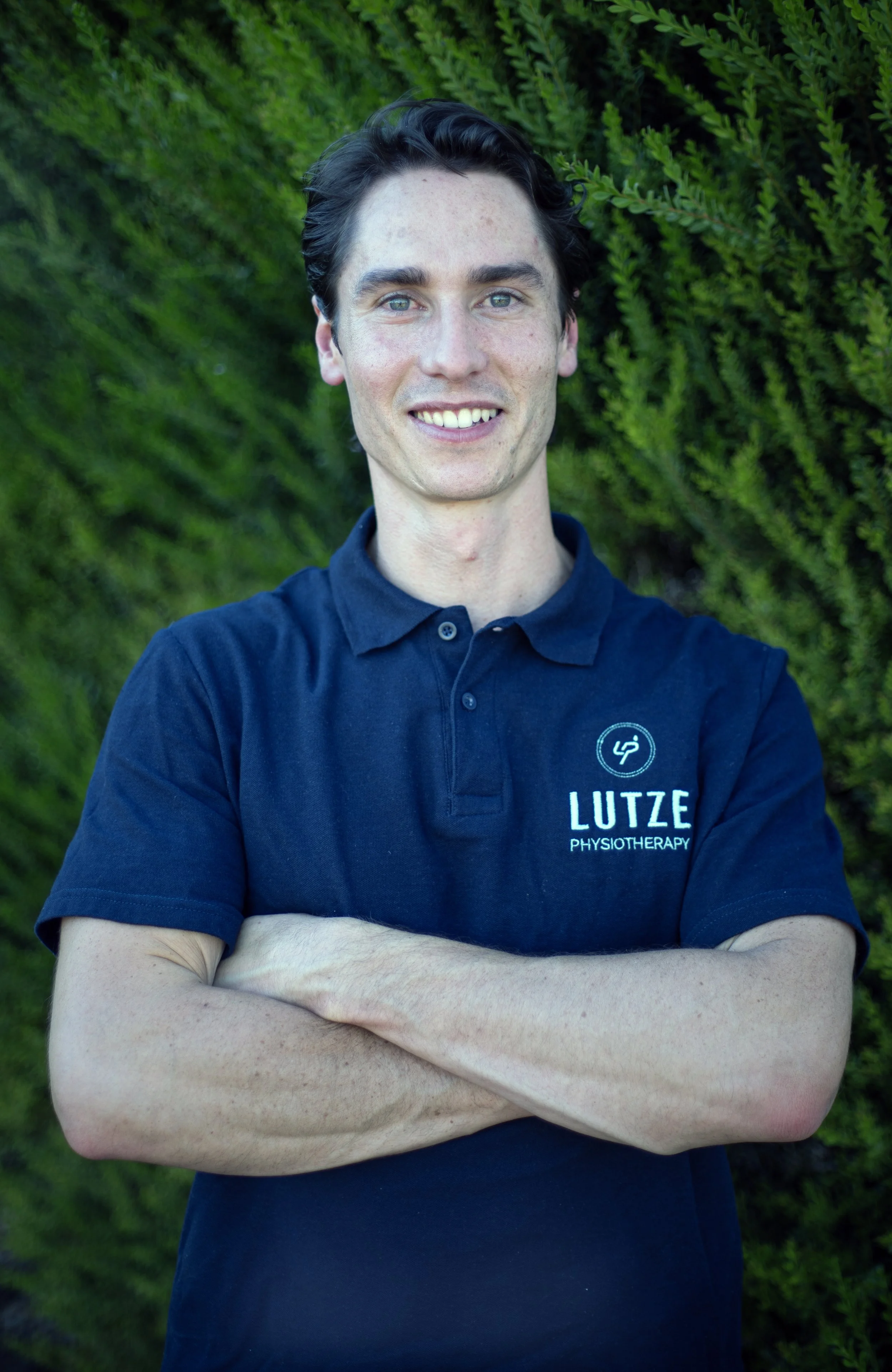 A young man with dark hair and fair skin, smiling and standing outdoors against a background of green foliage. He is wearing a navy blue polo shirt with the logo and name "LUTZE PHYSIOTHERAPY" on it, arms crossed.