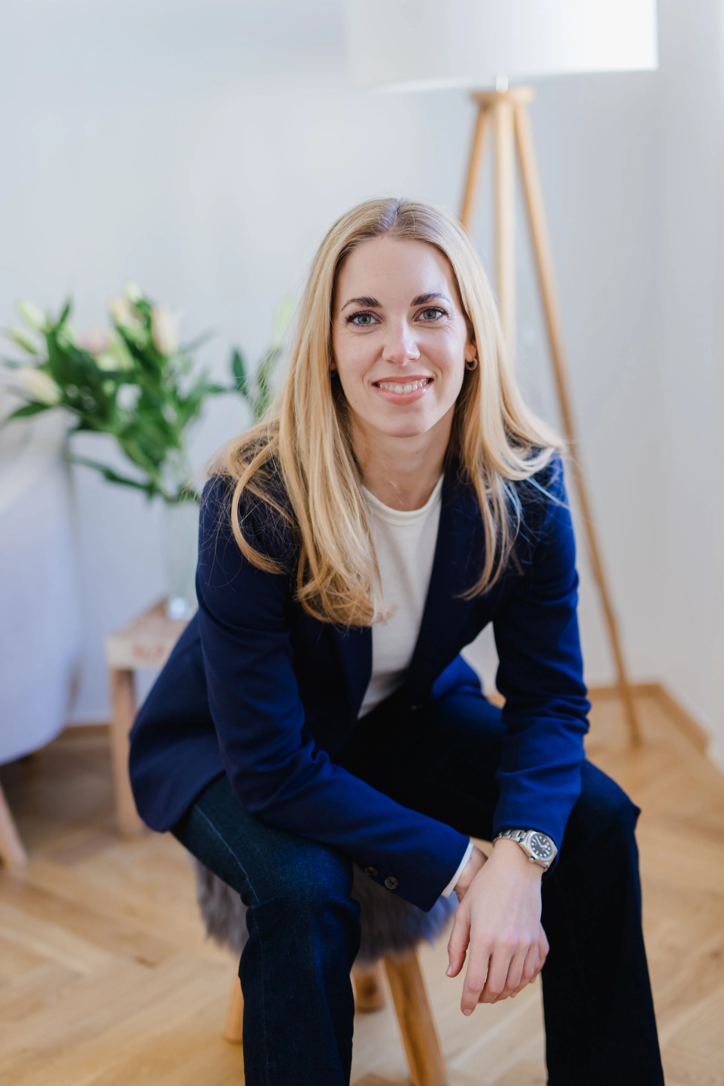 Junge Frau mit Blonden Haaren, dunkler Blazer und weißem T-Shirt, sitzt auf einem Stuhl in einem Raum mit Holzparkett, einem Lampenständer und einer Pflanze im Hintergrund.