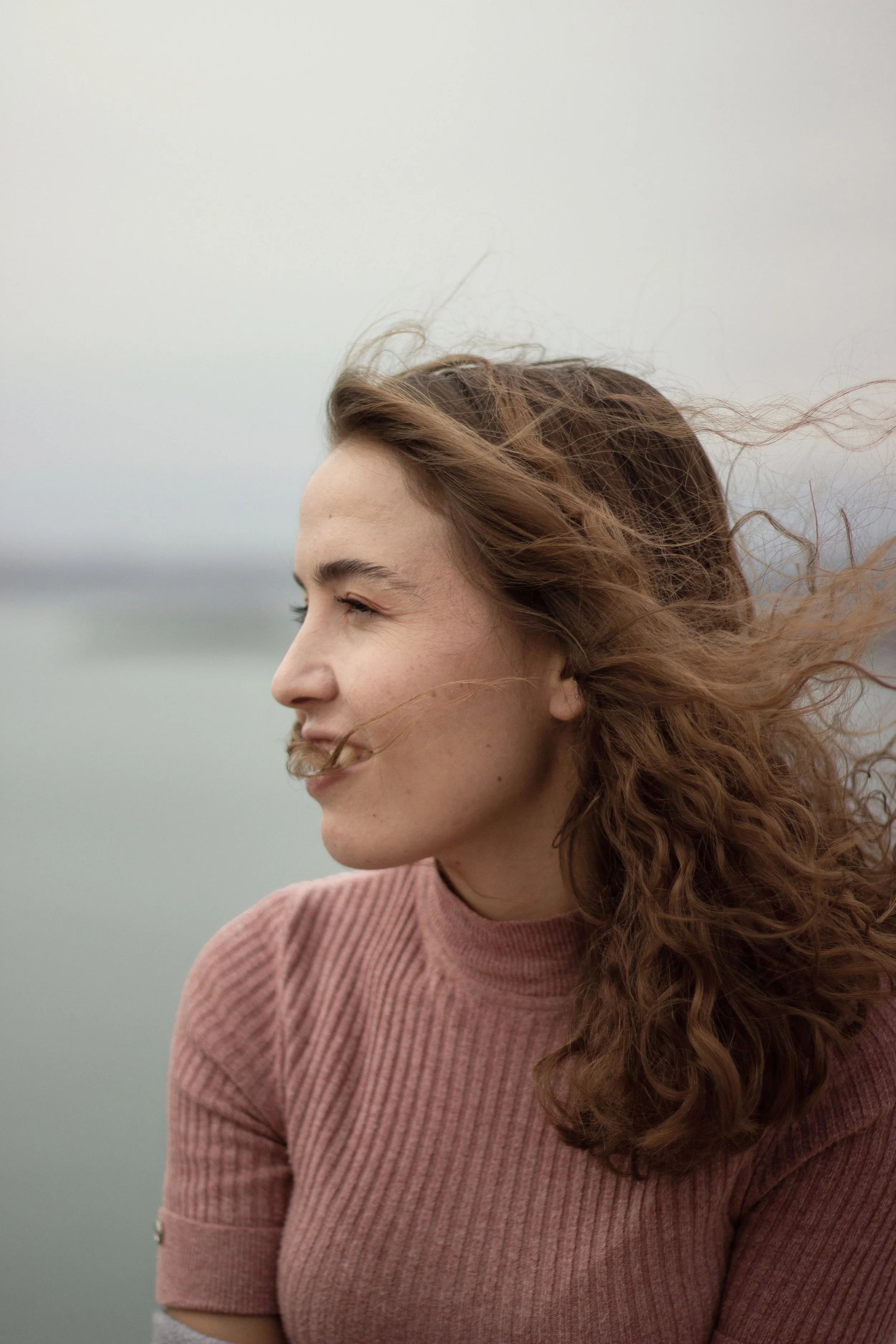 Frau im rosa Pullover blickt lächelnd zur Seite, während der Wind ihre Haare bewegt.