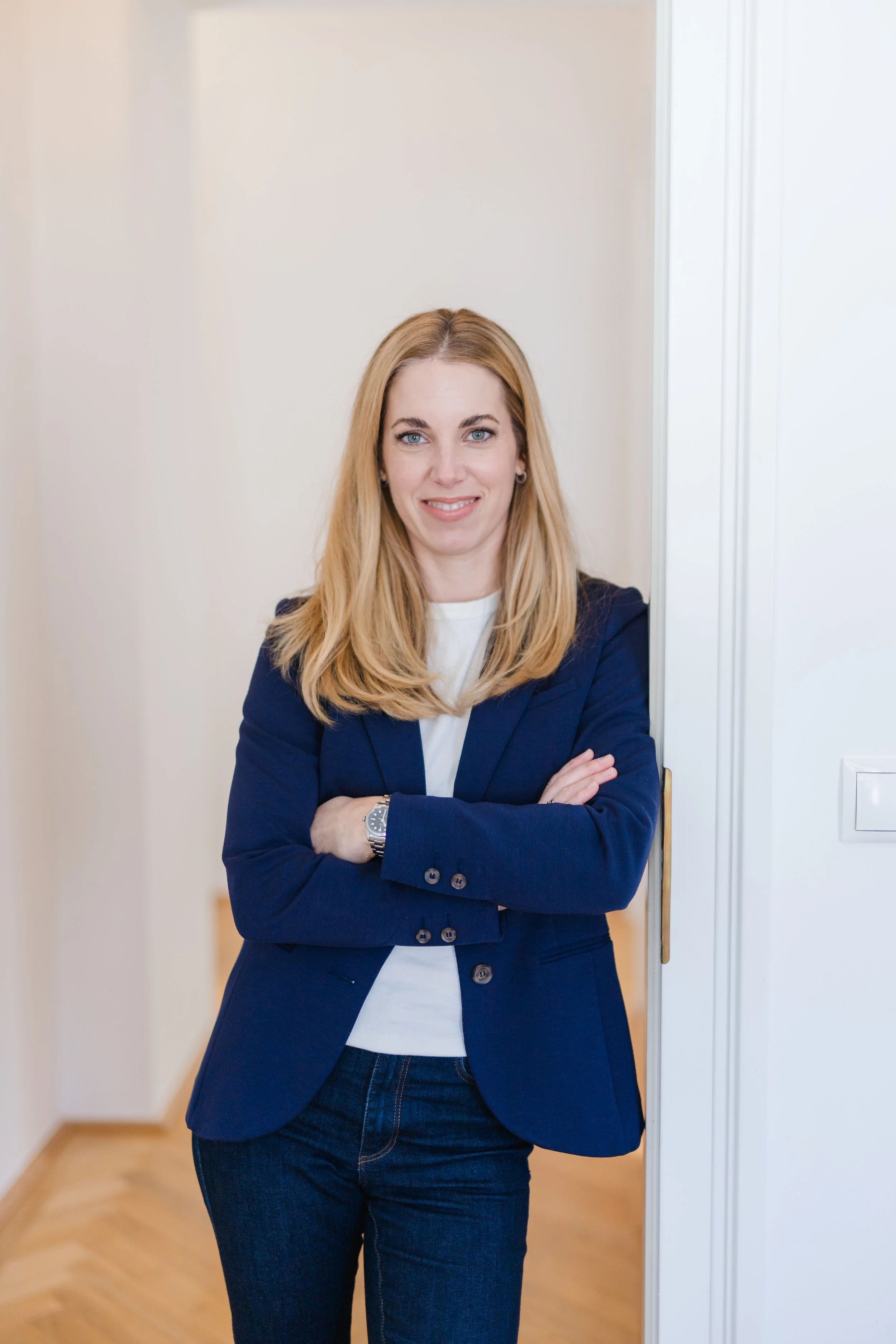 Eine Frau mit blonden Haaren, blauer Blazer und weißen Hemd steht mit verschränkten Armen vor einer weißen Tür.