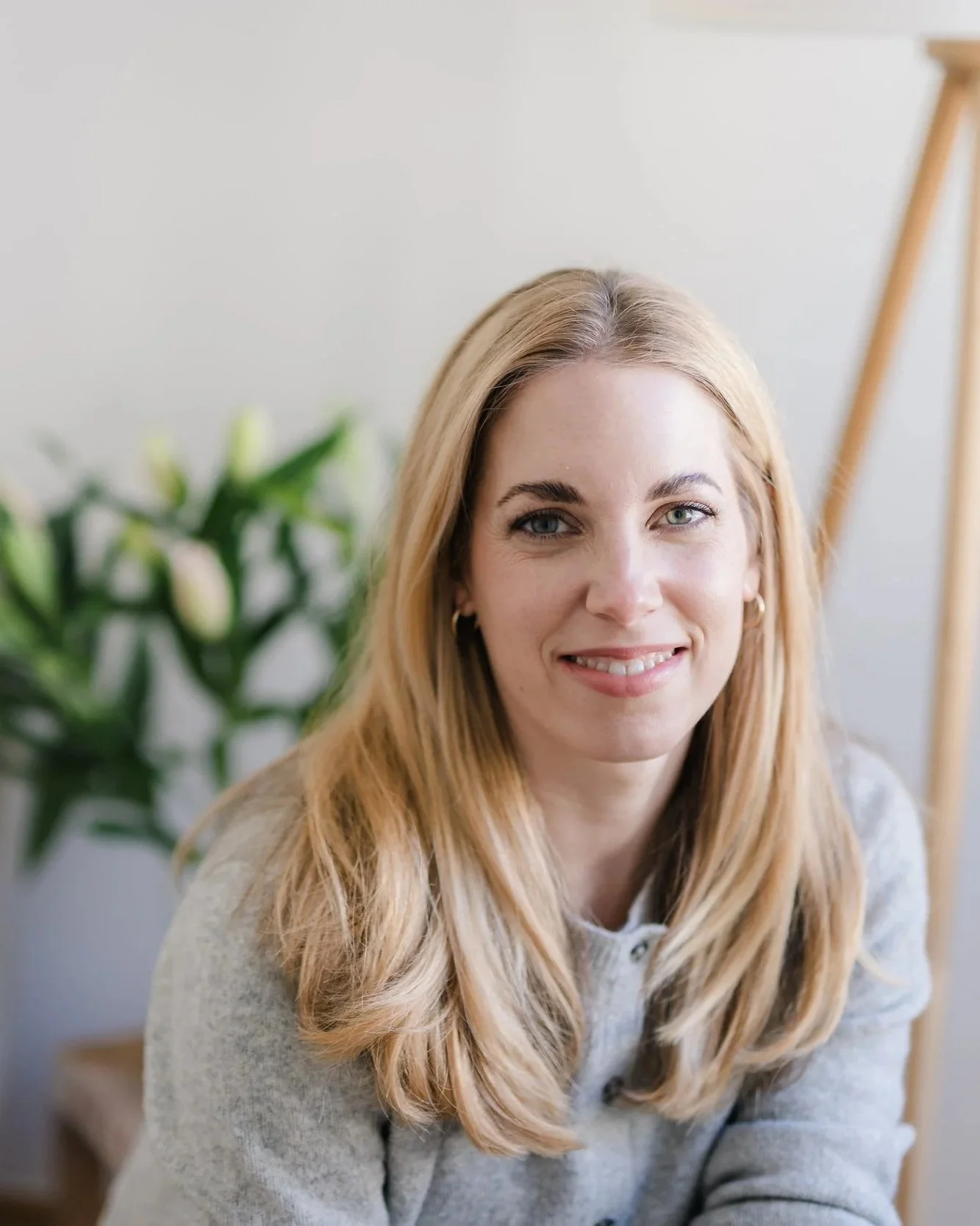 Eine Frau mit blonden Haaren, lächelnd, im Wohnzimmer mit Pflanze im Hintergrund.