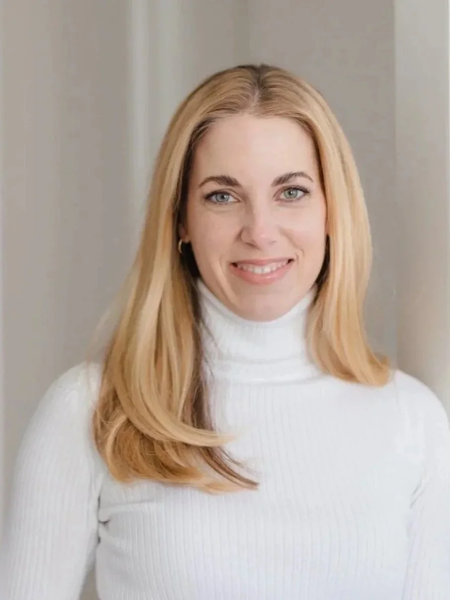 Lächelnde Frau mit langen blondem Haar, trägt weißen Rollkragenpullover, im Hintergrund eine weiße Wand.