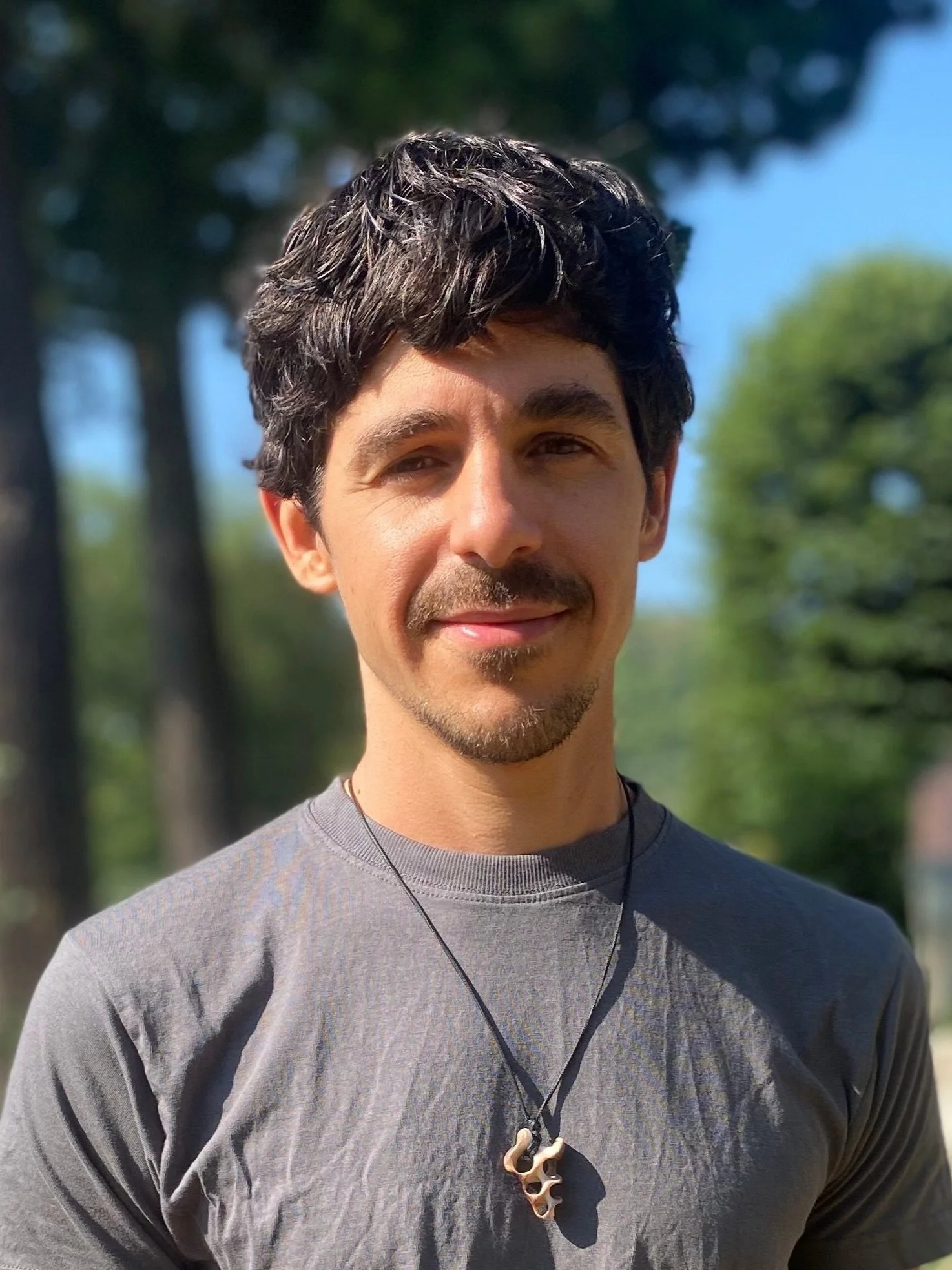 A young man with dark, wavy hair and a mustache is smiling outdoors on a sunny day, wearing a gray athletic shirt and a necklace with a pendant, with blurred trees in the background.