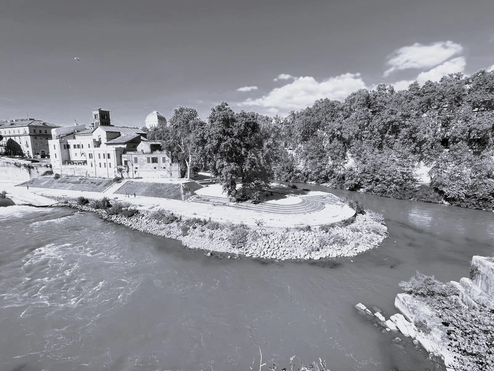Black and white photo of a river with a small peninsula featuring a permanent concert area, trees along the riverbank, and buildings in the background.