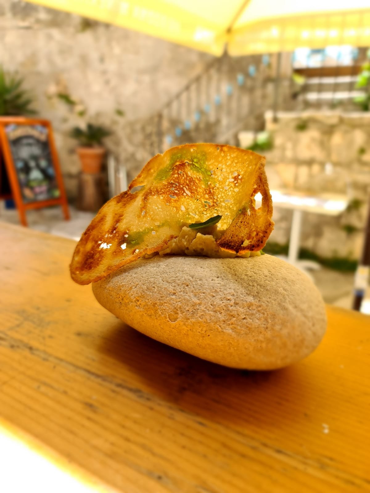 A crispy golden-brown slice of vegetable omelette resting on a smooth round stone, placed on a wooden surface, with an outdoor patio and stairs in the background.