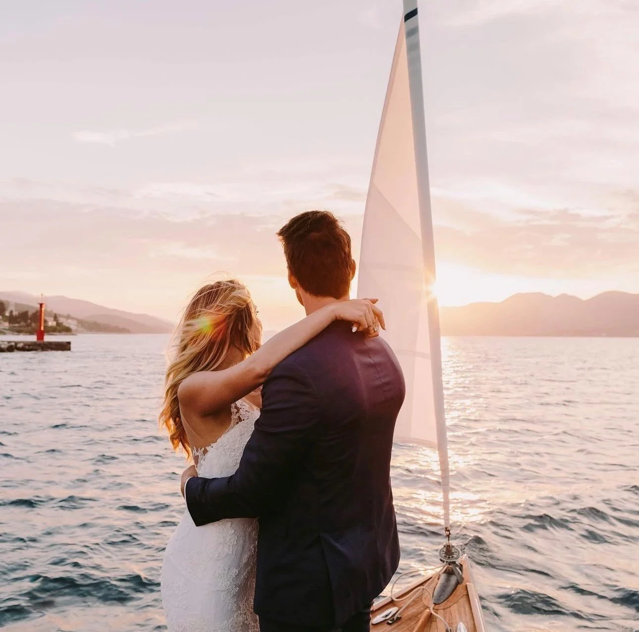 A couple, dressed in wedding attire, embracing on a boat at sunset with mountains in the background and a sailboat nearby.