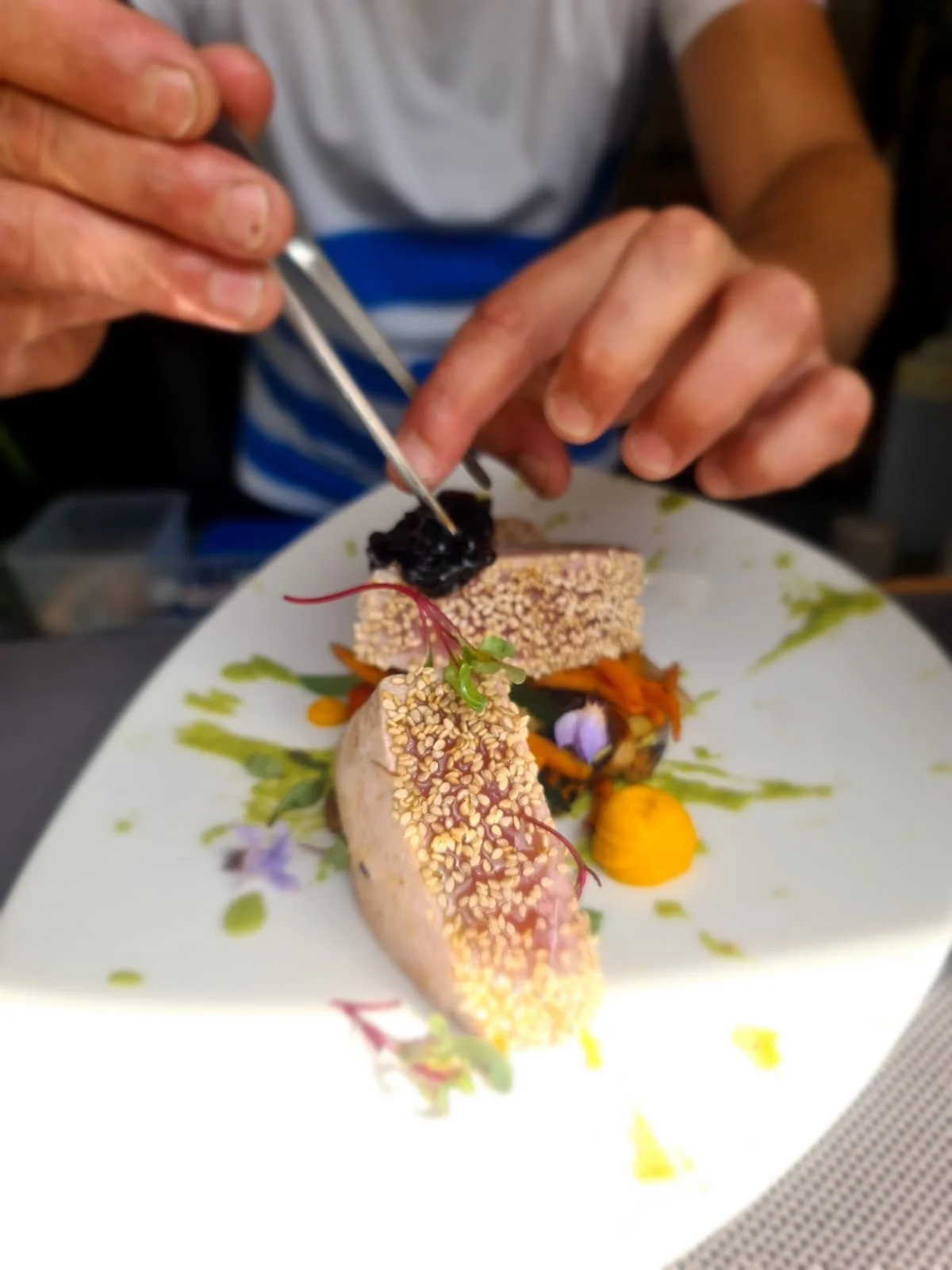 Person adding a dark sauce or garnish to a plated dish with sesame-crusted meat, vegetables, and edible flowers.