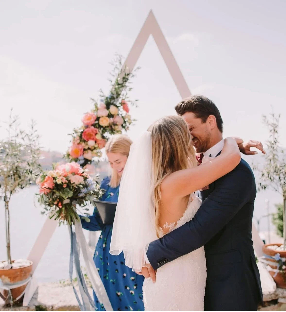 Bride and groom embrace at their wedding ceremony outdoors, with an officiant and floral decorations in the background.