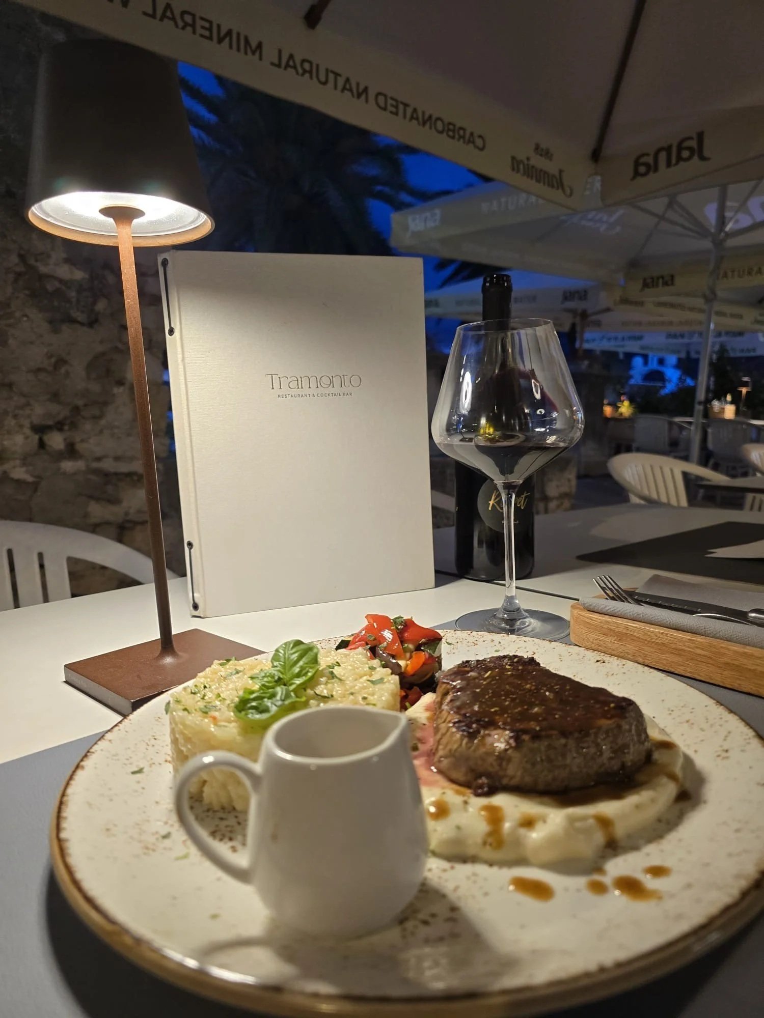 A plate of steak with mashed potatoes, a side of vegetables, a small pitcher of sauce, a glass of red wine, a menu, and a bottle of wine on a table at a restaurant outdoor patio during evening.