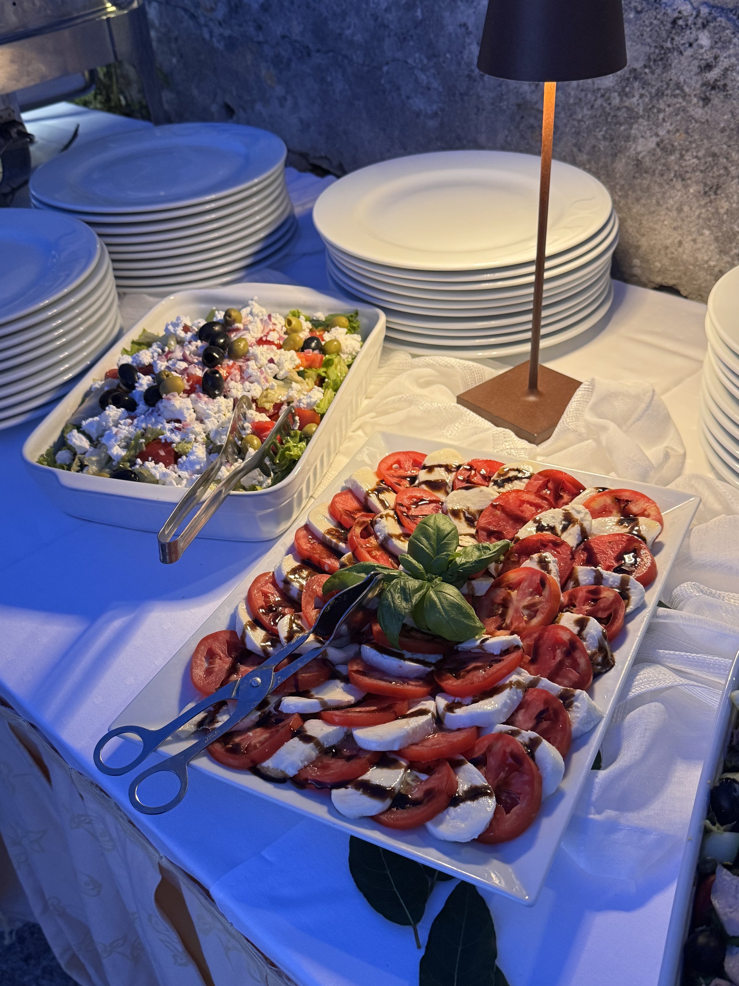 Buffet table with plates, a tomato and mozzarella salad with basil, and a mixed salad with olives, feta cheese, and vegetables, under a black lamp.