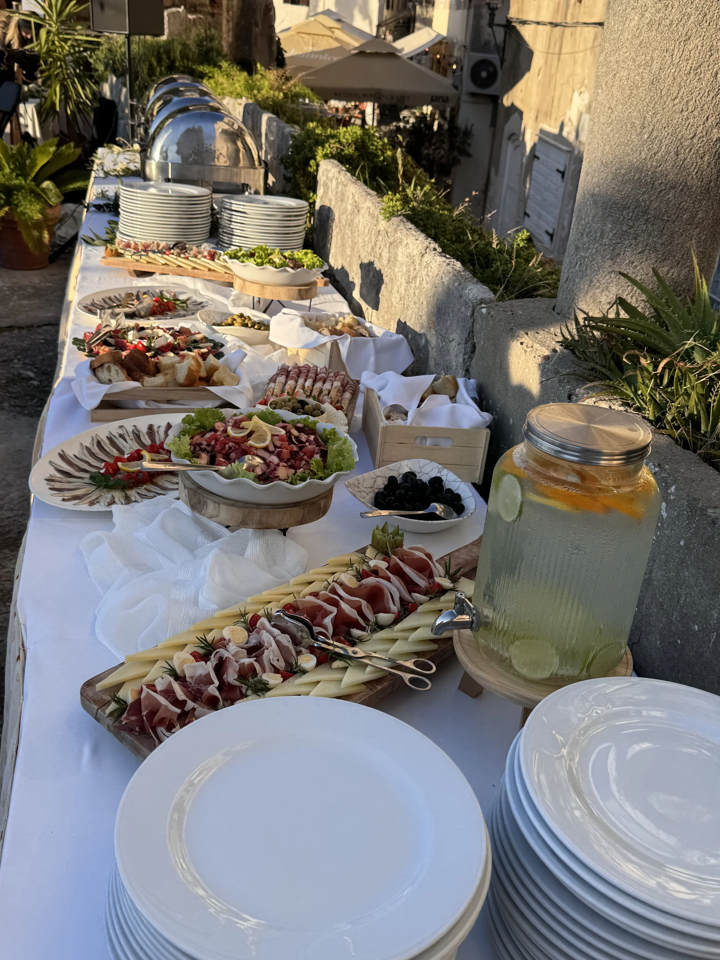 Buffet table with assorted appetizers, cold cuts, cheeses, salads, and a large jar of infused water, set outdoors with plates and utensils ready for guests.