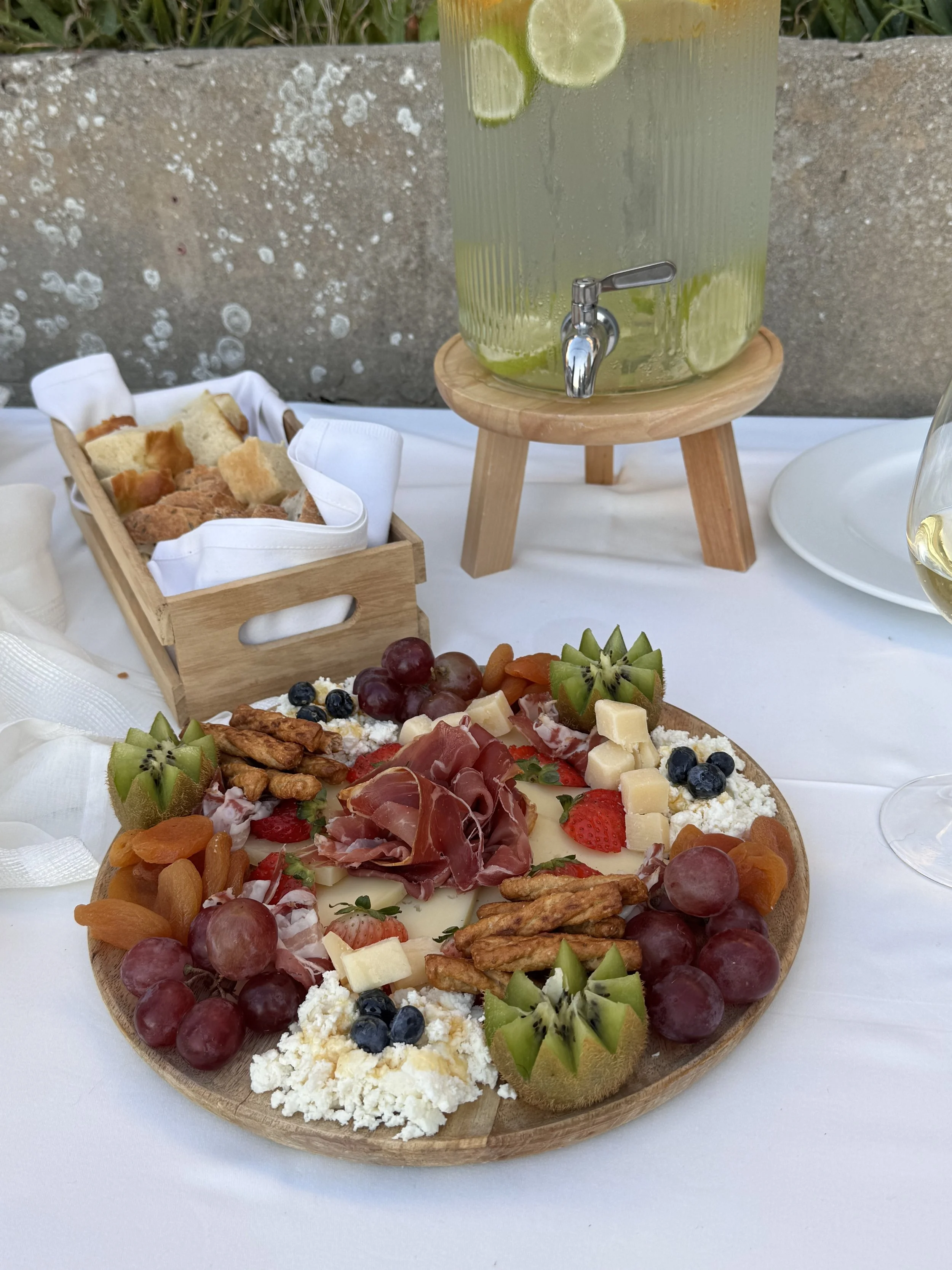 A charcuterie platter with grapes, strawberries, blueberries, kiwifruit, cheese, honey, and prosciutto, served on a wooden board. A glass of white wine and a pitcher of lime-infused water are also on the table.