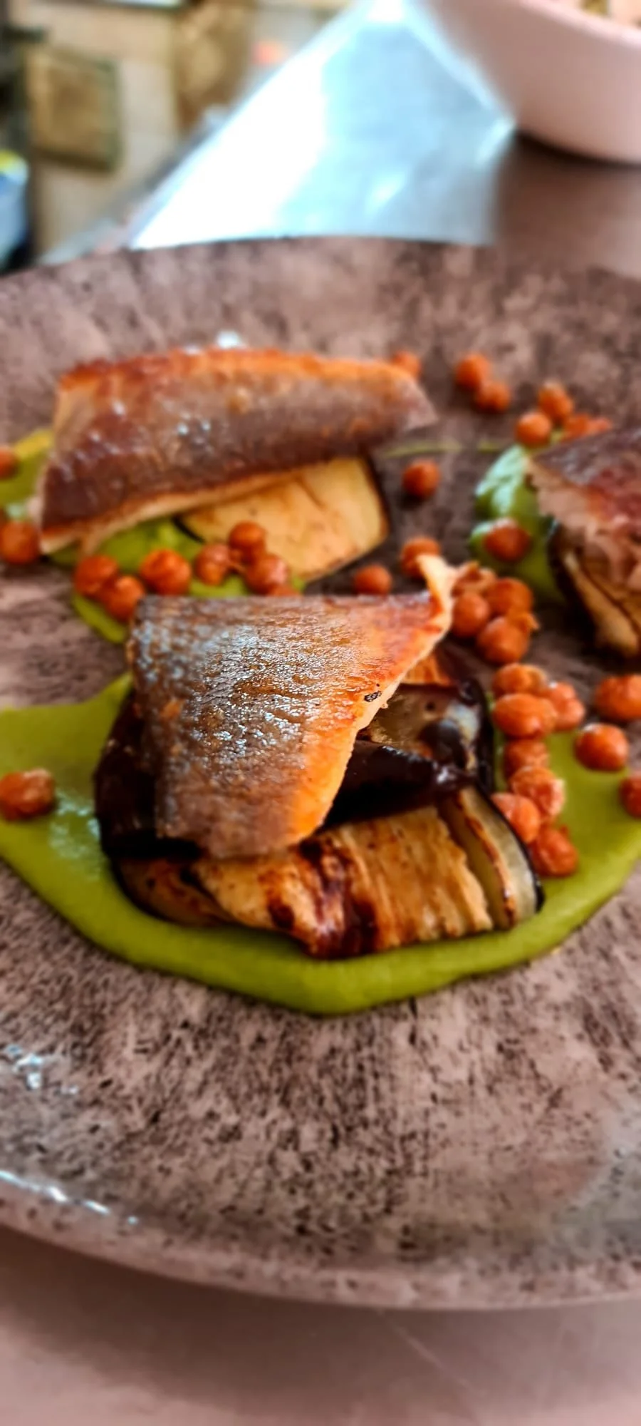 Close-up of grilled fish with crispy skin served with grilled eggplant and chickpeas on a green sauce on a brown plate.