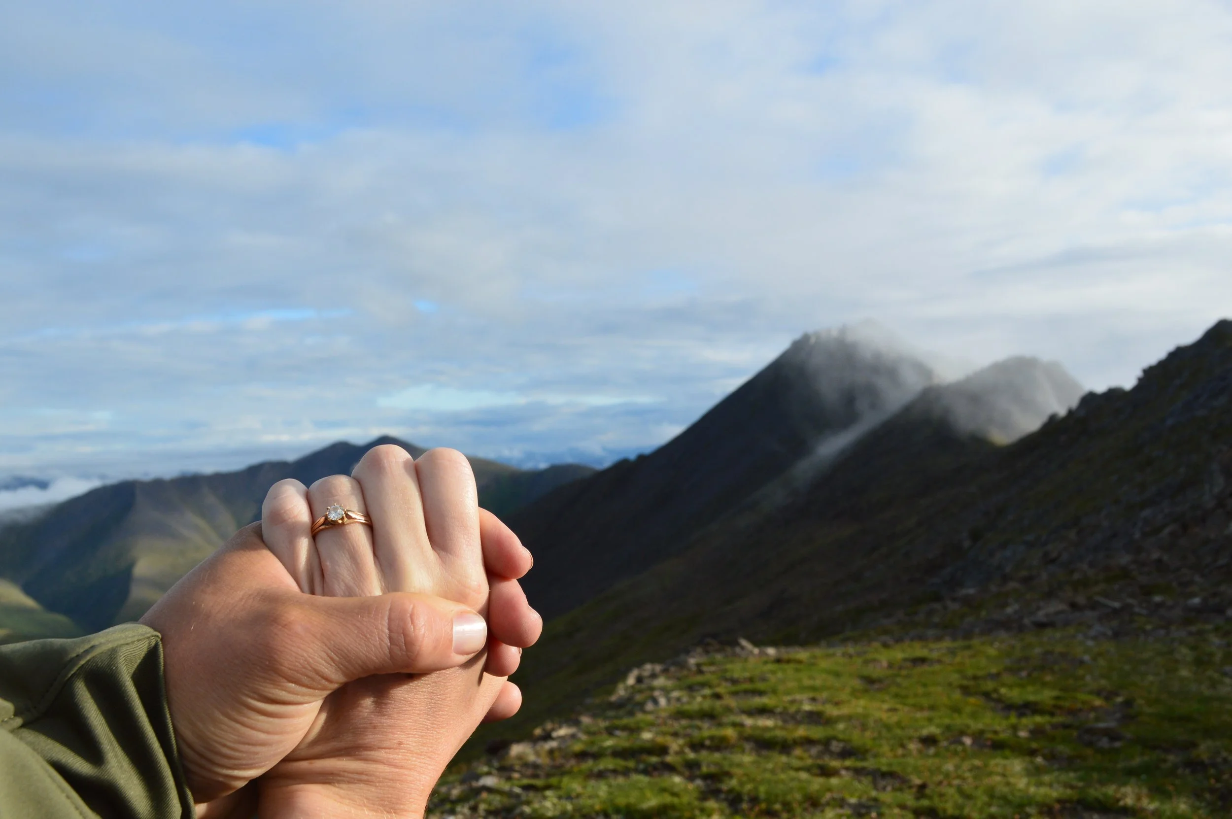 Zwei Hände, die einander halten, mit einem Verlobungsring auf dem Finger, vor einer Berglandschaft mit Wolken am Himmel.