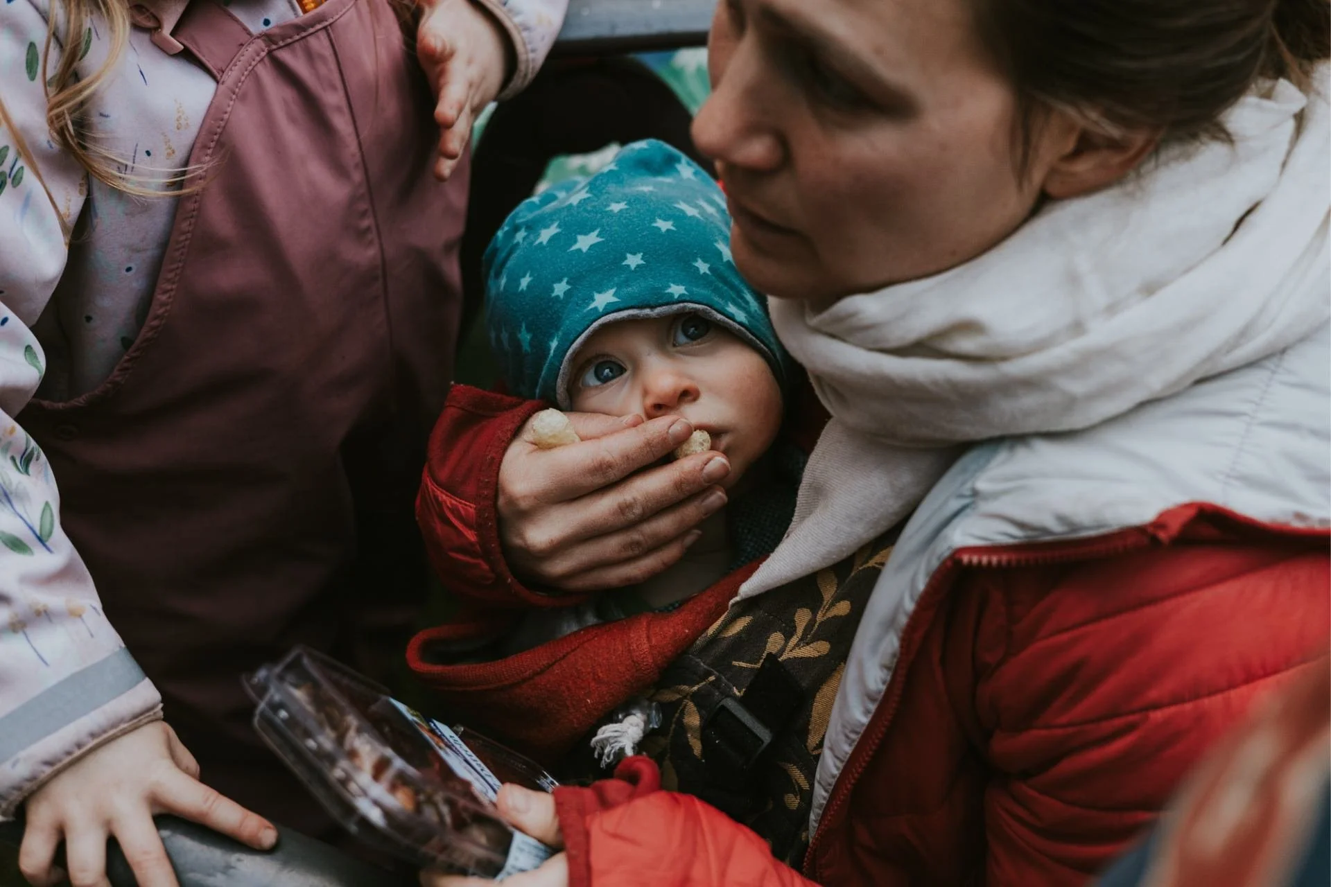 Ein Baby mit blauen Augen, das einen hellblauen Mütze mit Sternen trägt, wird von der Mutter gehalten, die es liebevoll füttert und gleichzeitig mit dem großen Kind spricht. Das Baby schaut mit großen Augen direkt zur Mutter.