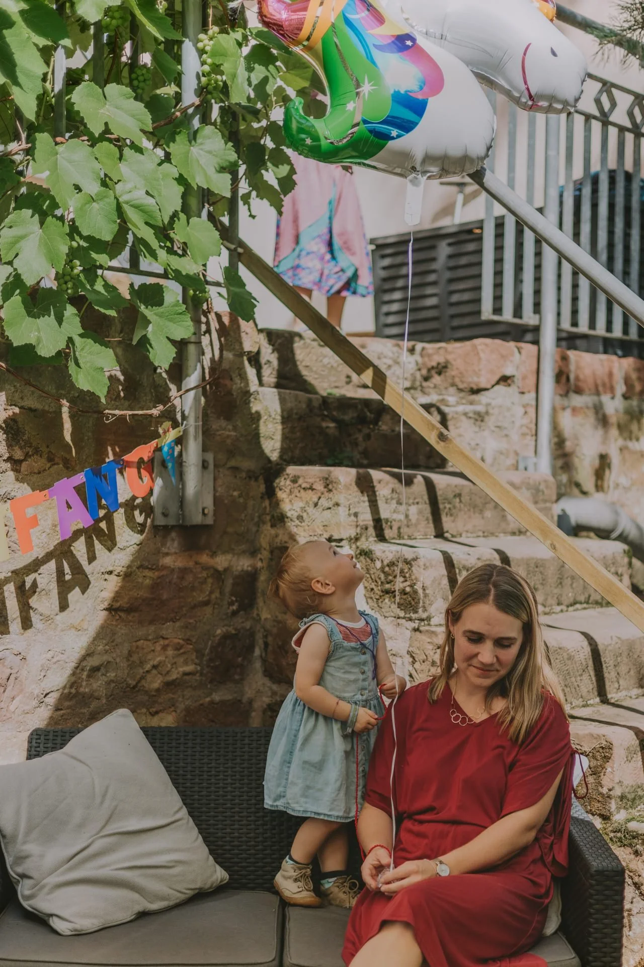 Kleines Kind steht neben Mutter auf dem Sofa im Garten und blickt fröhlich zum Ballon beim Einschulung feiern in der Pfalz.