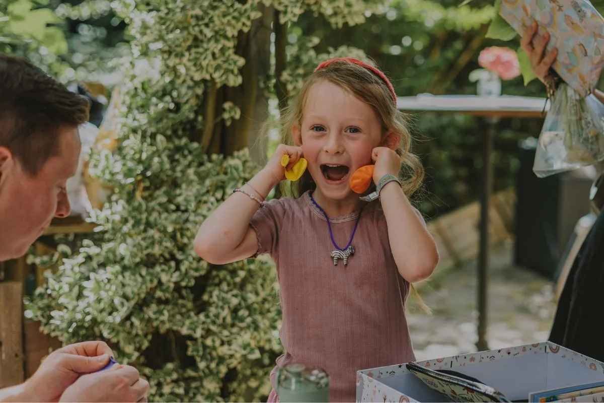 Mädchen hält sich lachend die Ohren zu während Papa Luftballon Geräusche macht bei Einschulungsfeier im Garten.