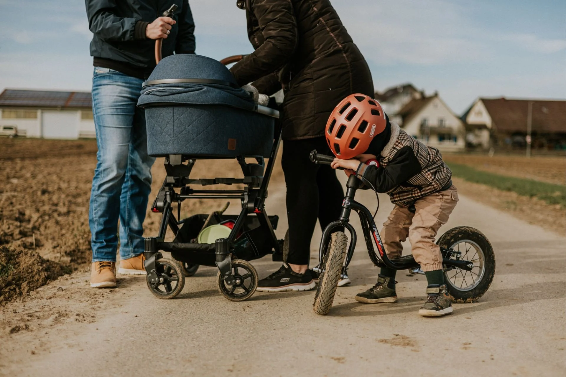 Eine Familie bei einem Spaziergang auf einem Feldweg, Kind mit Fahrradhelm wartet genervt und lehnt seinen Kopf an den Laufrad-Lenker, während die Erwachsenen versuchen das Baby zu beruhigen.
