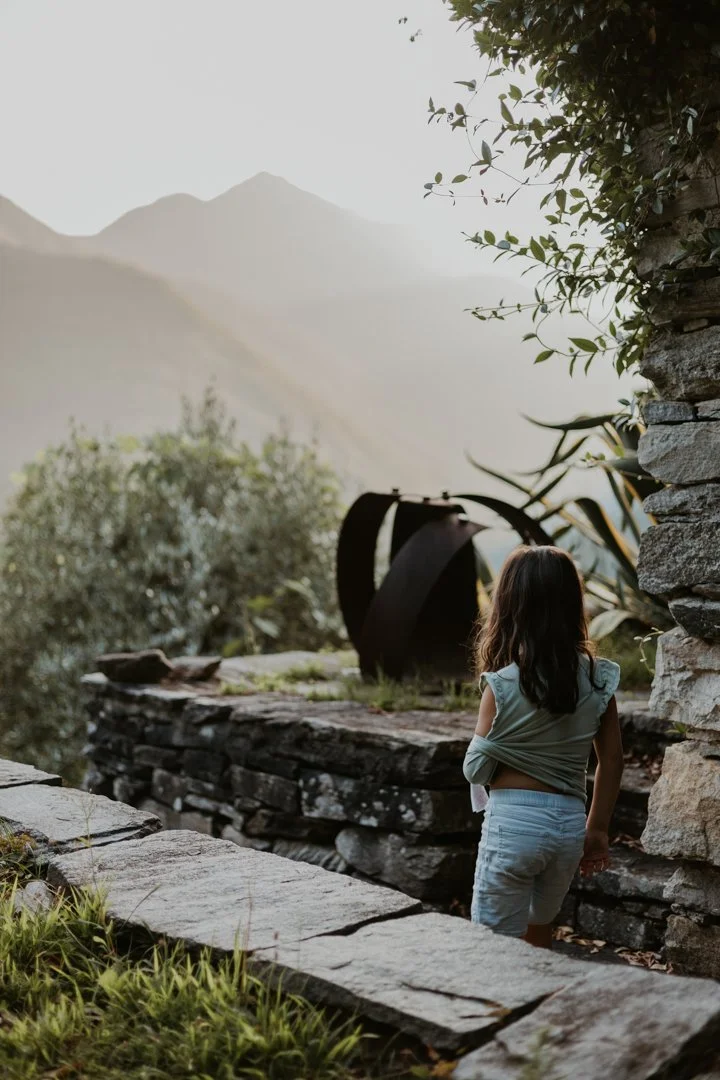 Ein junges Mädchen mit braunen Haaren, das auf einer alten Steinterrasse steht und in die Ferne blickt, umgeben von Natur mit Bergen im Hintergrund.