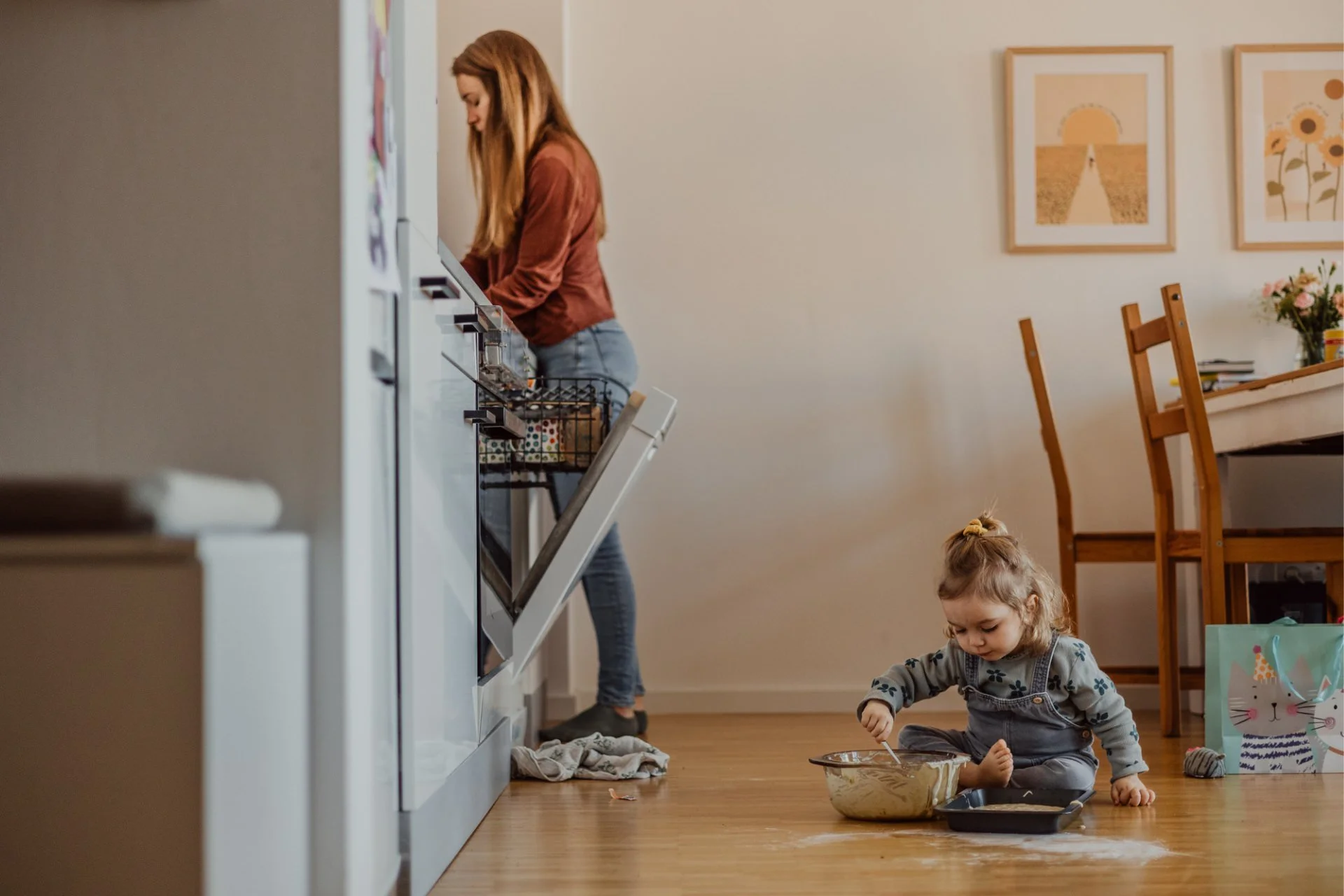 Eine junge Mutter ist in der Küche tätig, während ein kleines Mädchen auf dem Boden sitzt und in einer Schüssel mit Teig rührt.