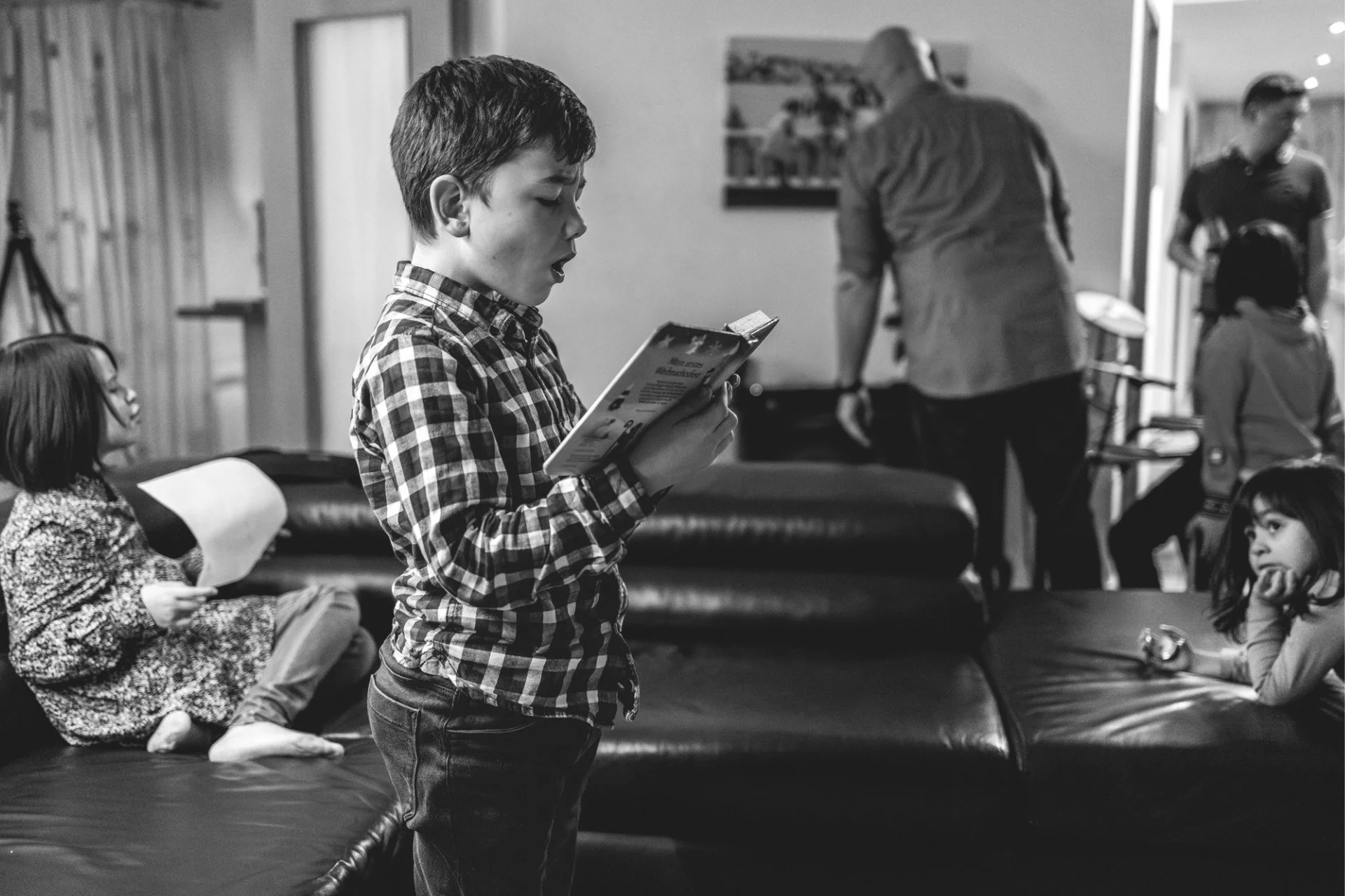 Junge trägt etwas aus einem Buch vor in einem Wohnzimmer voller Menschen, die durcheinander laufen. Ein Kind beobachtet ihn fasziniert.