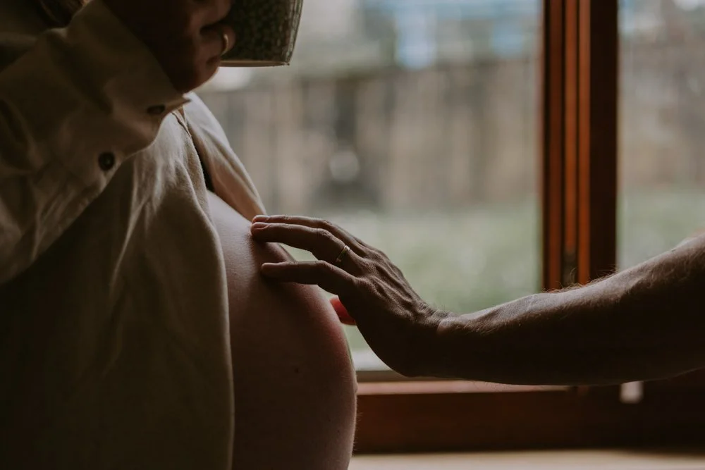 Eine schwangere Frau wird von einer Hand am Bauch gestreichelt, im Hintergrund ein Fenster mit Blick nach draußen.