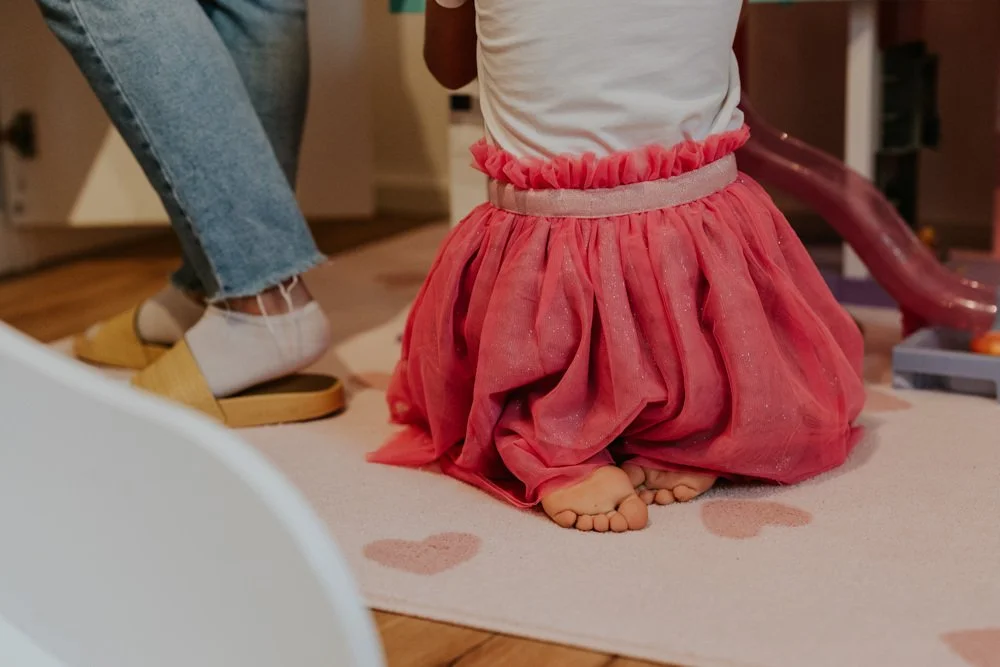 Ein barfüßiges Mädchen in einem pinken Tüllrock, das auf den Knien auf einem Teppich sitzt.