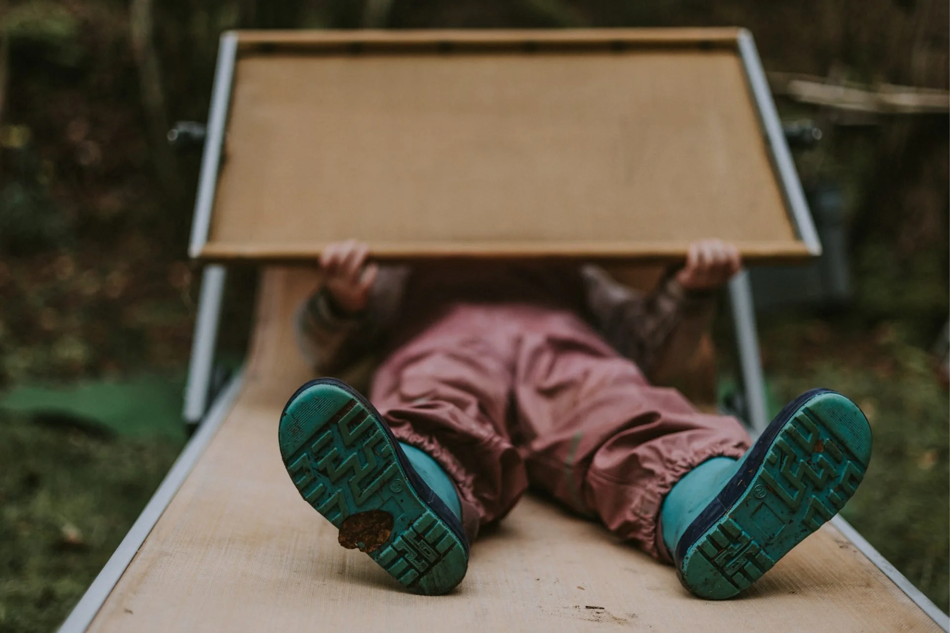 Ein Kind liegt auf einem Sonnenstuhl mit Blick nach oben, die Füße in den Vordergrund mit blauen Gummistiefeln, im Hintergrund ist ein Garten sichtbar.