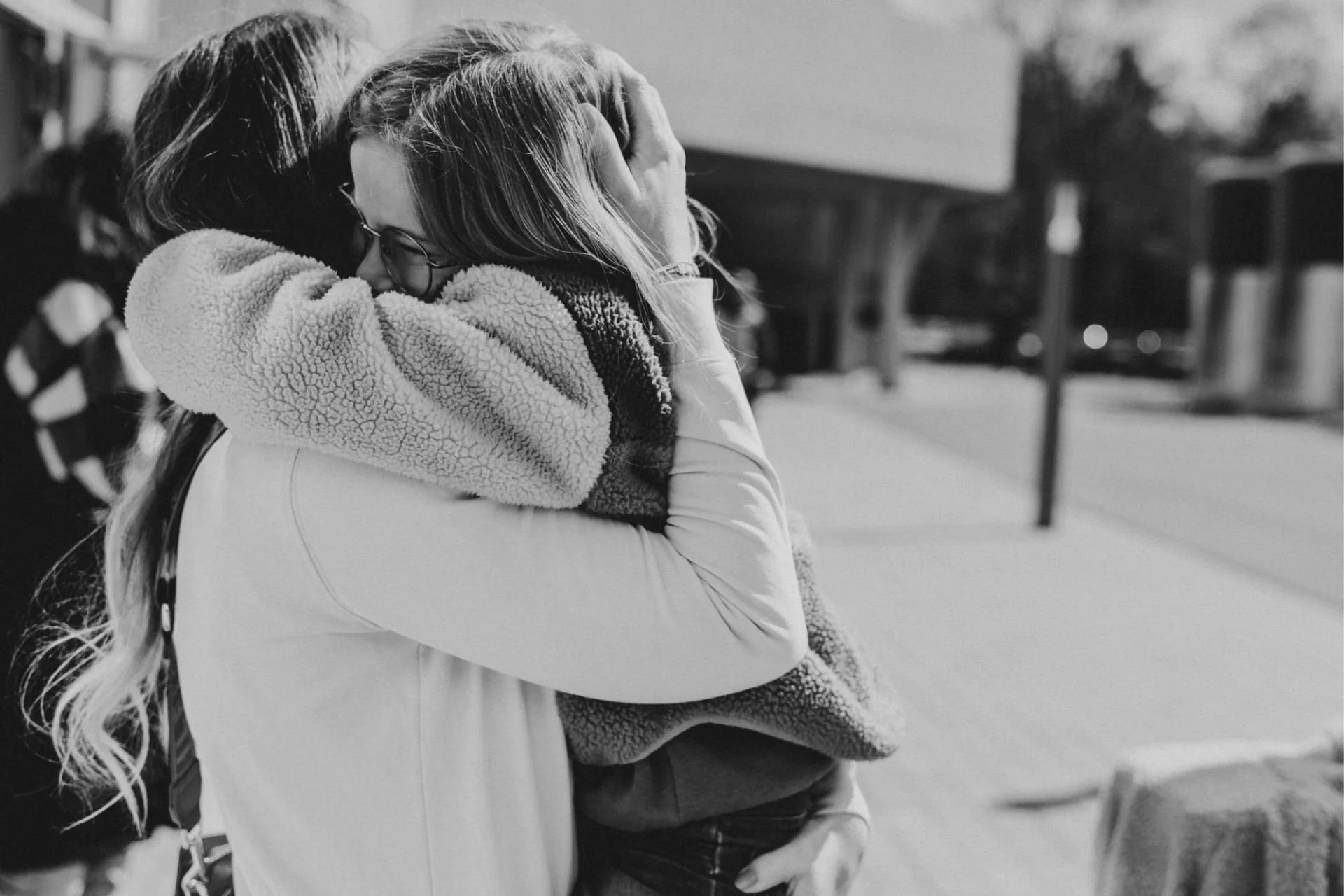 Eine Mama und ihre kleine Tochter umarmen sich im Freien, die Tochter trägt eine Brille und eine warme Jacke.
