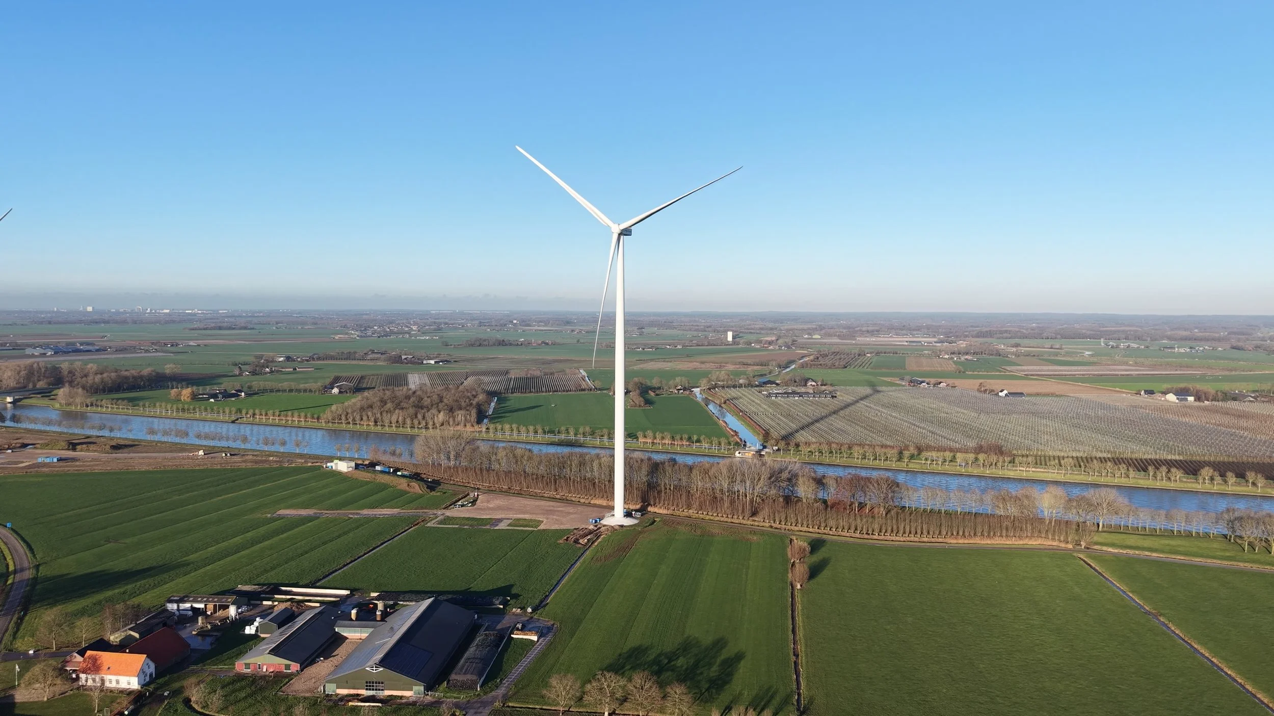 Luchtfoto van een landschap met een grote windturbine in het midden, omgeven door groene velden, rivieren en weilanden, onder een heldere blauwe lucht.