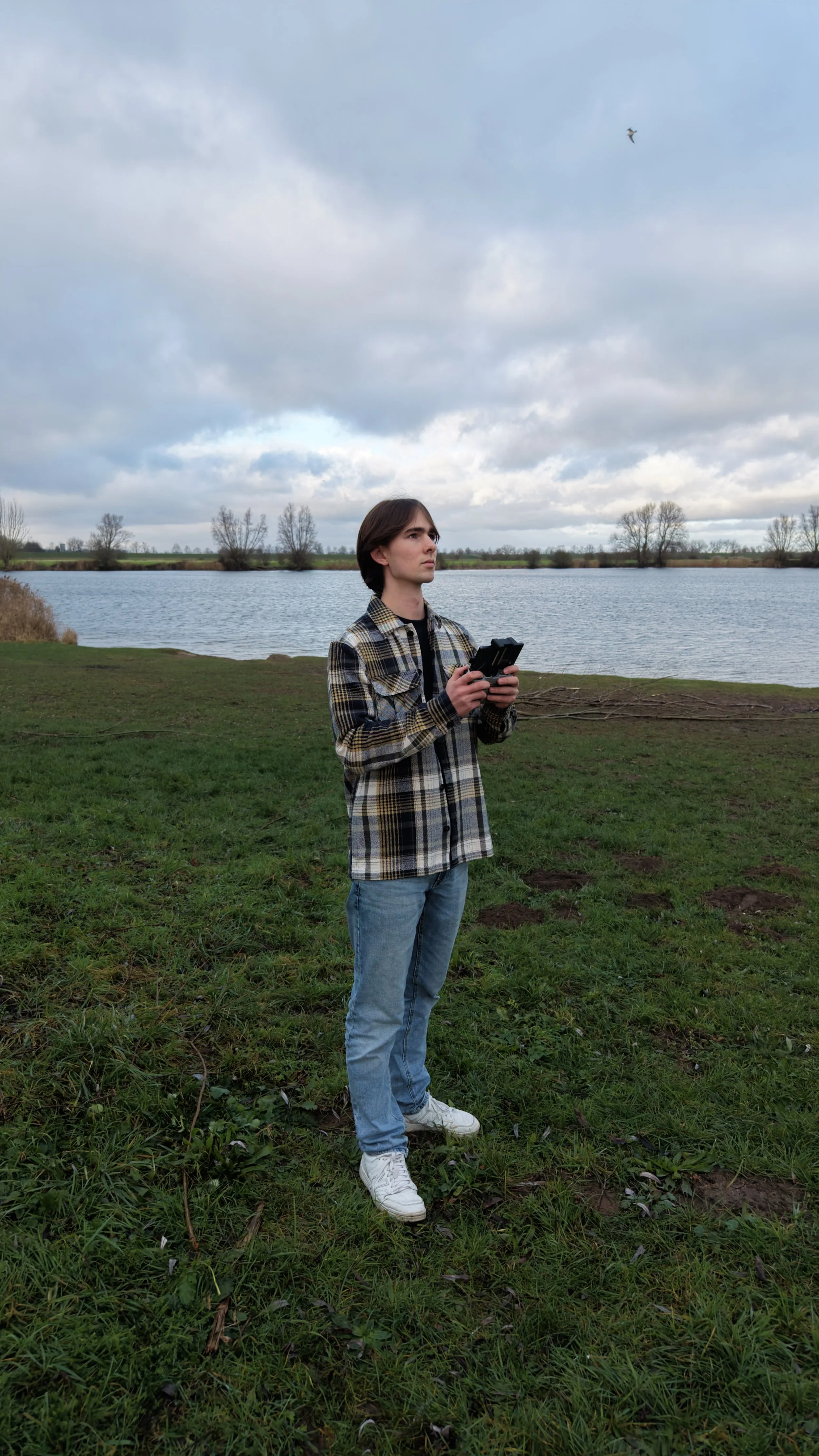 Jongen met lange bruin haar, spijkerbroek en een plaid overhemd, staand op gras bij een meer, die een afstand bedient met een drone onder een bewolkte hemel.