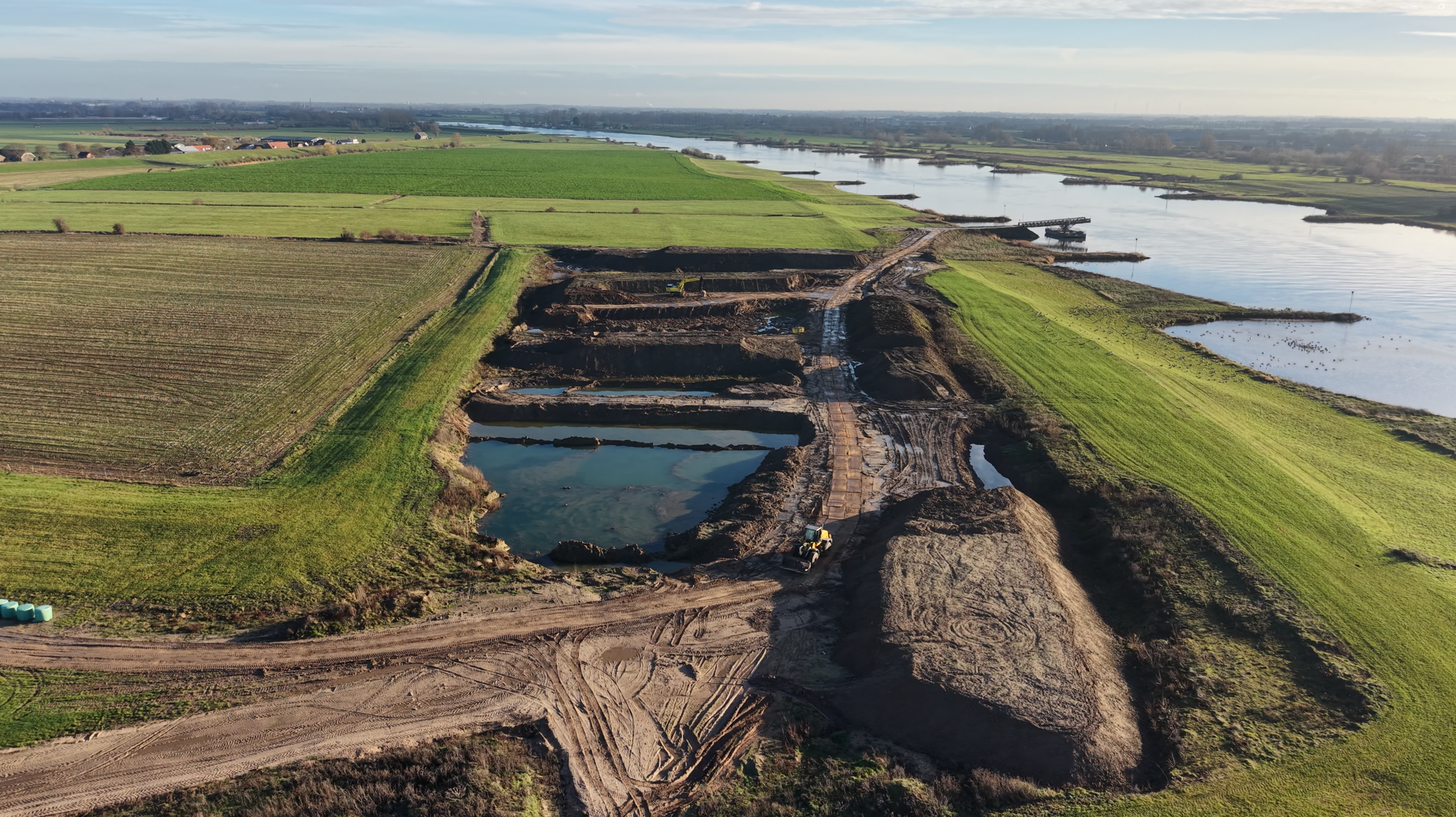 Luchtfoto van een bouwplaats naast een rivier, met graafmachines, waterbassins en uitgestrekte groene landbouwvelden.