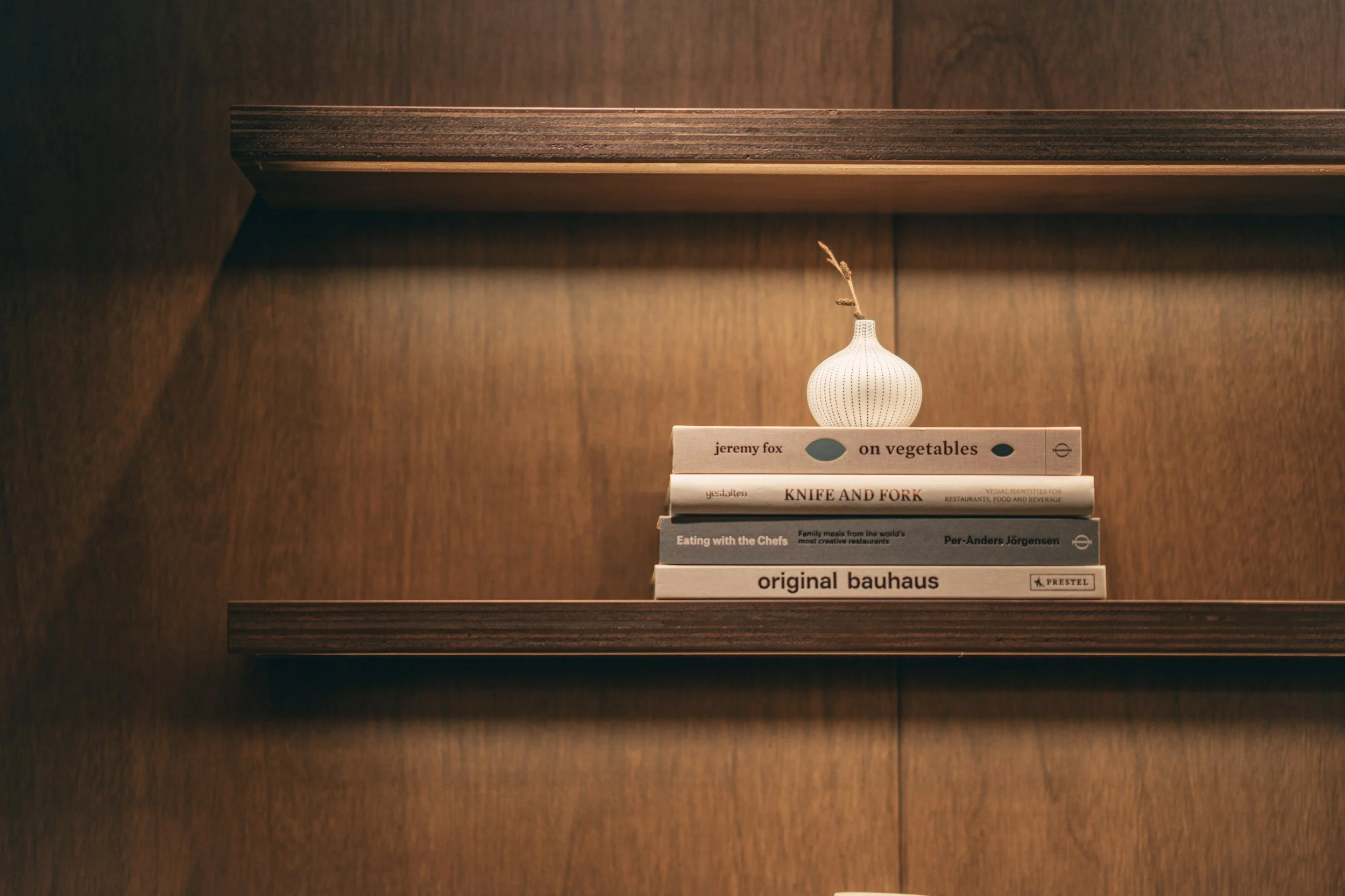 A small white vase with a textured surface and a single dried flower stem inside it, sitting on a stack of five cookbooks on a wooden shelf with a wood panel background.