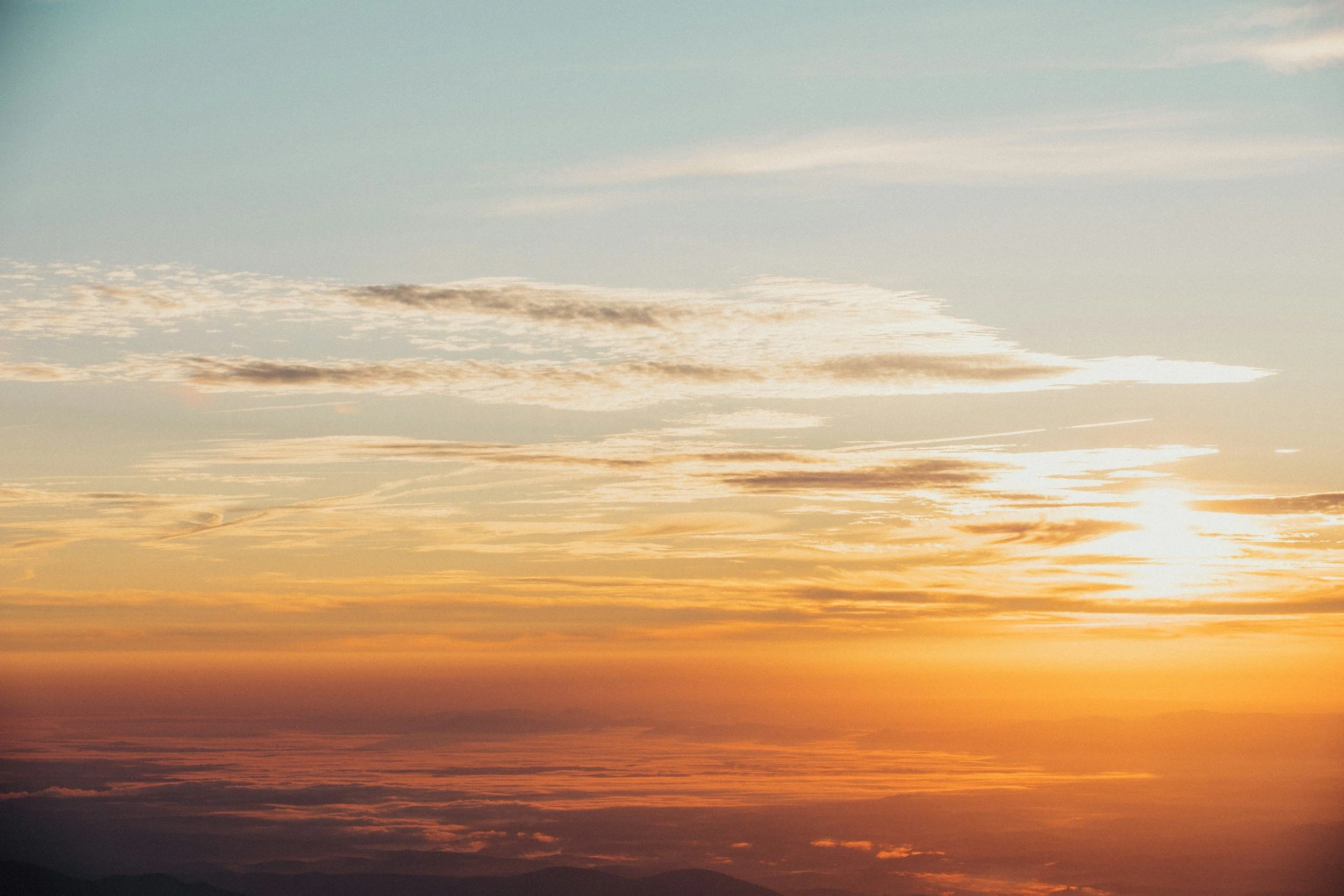 A sunset sky with clouds, showing warm orange and yellow hues near the horizon, transitioning to cooler blue tones higher up.