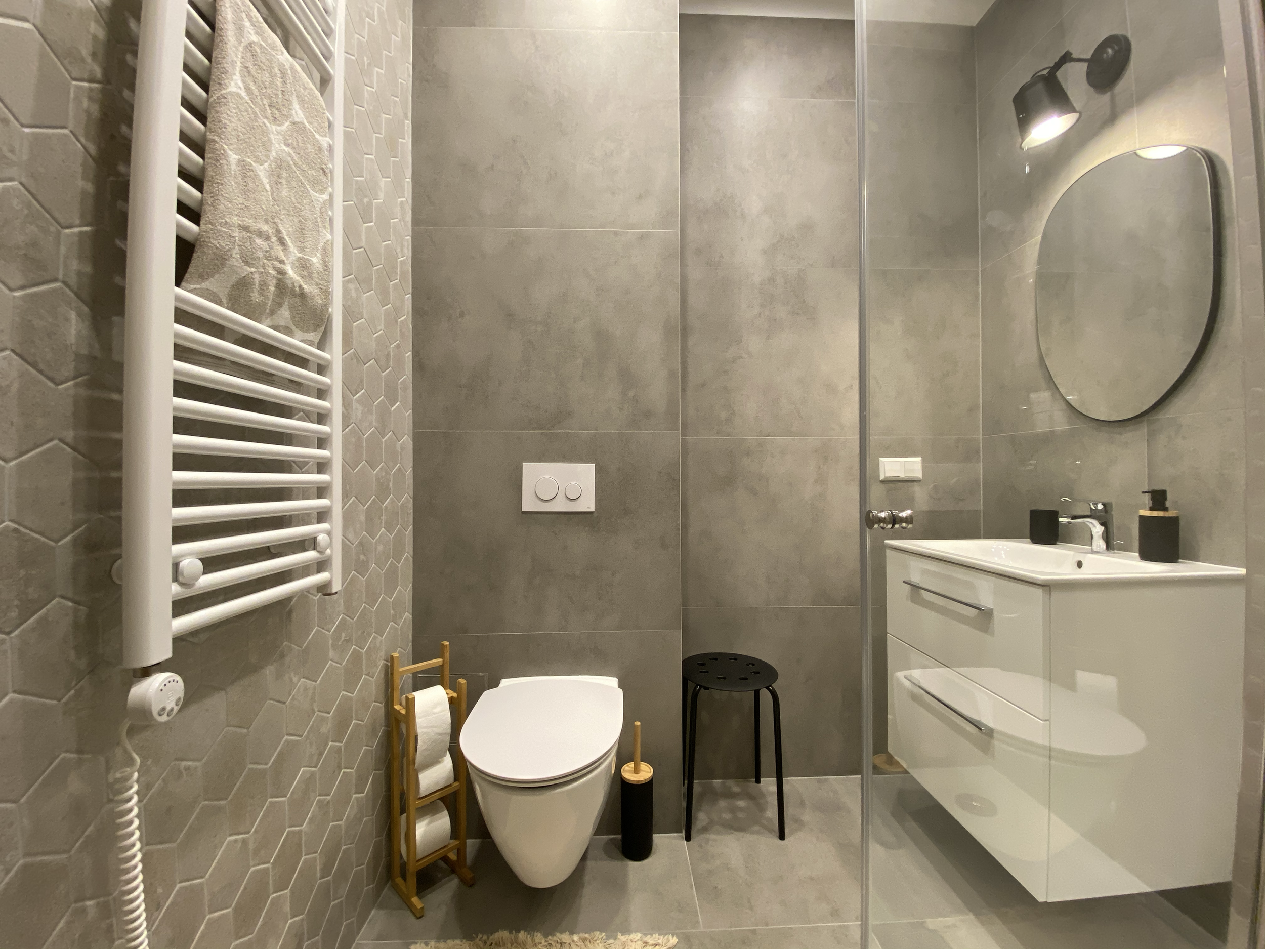 Modern bathroom with hexagonal tiles and gray wall tiles. Features include wall-mounted toilet, white vanity with sink, round mirror, black shower lamp, black soap and toothbrush holders, towel radiator, small black stool, toilet paper holder, and toilet brush.
