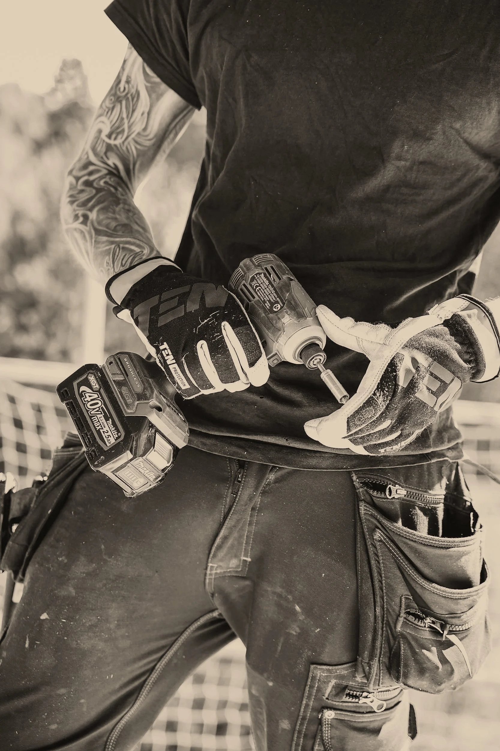 A person wearing gloves and work clothes is holding a cordless drill, with a retractable tape measure hanging from their pocket, on a construction site.