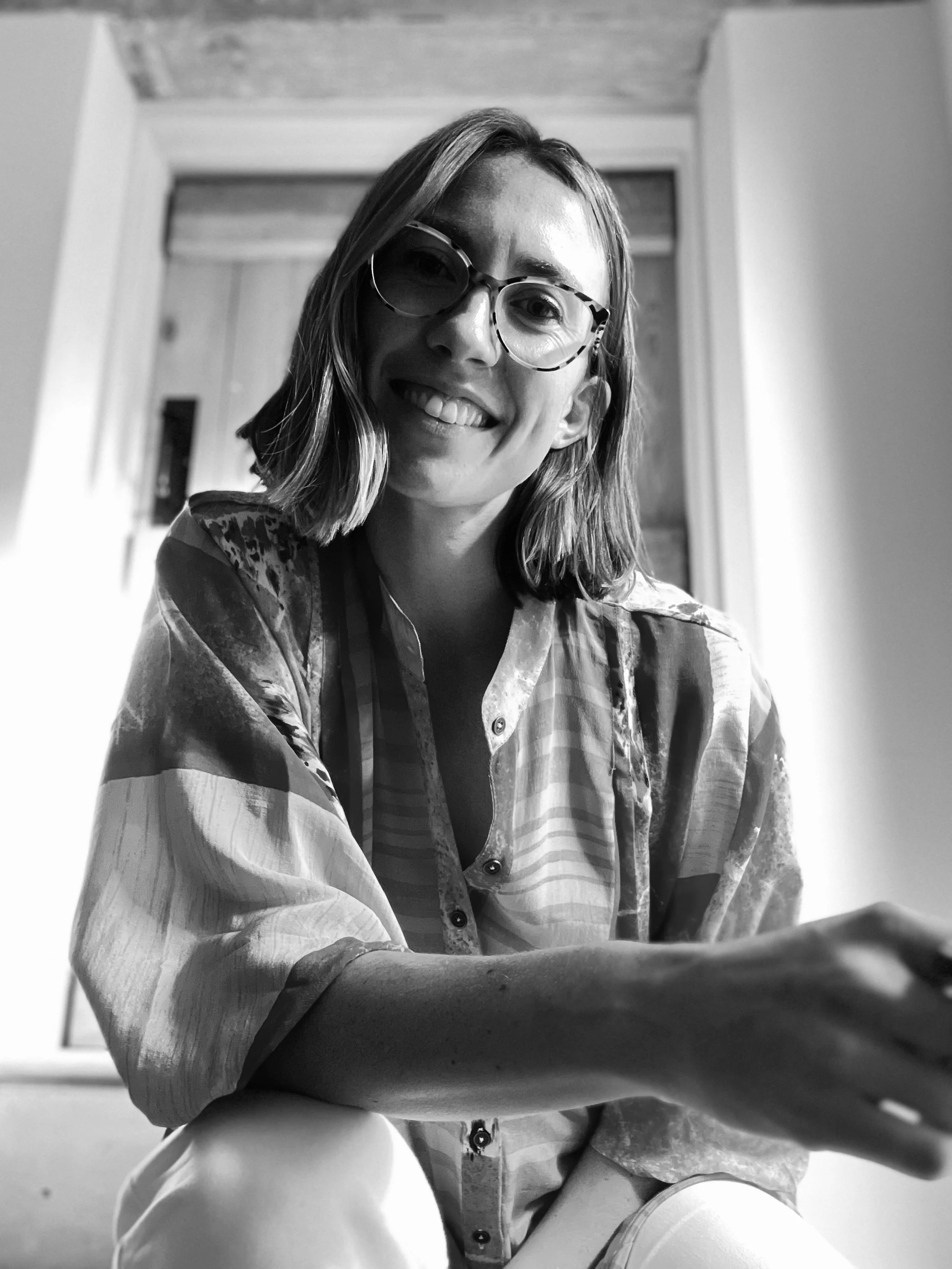A black and white photo of a woman smiling, wearing large glasses and a patterned shirt, seated and looking at the camera with her hand resting on a surface.