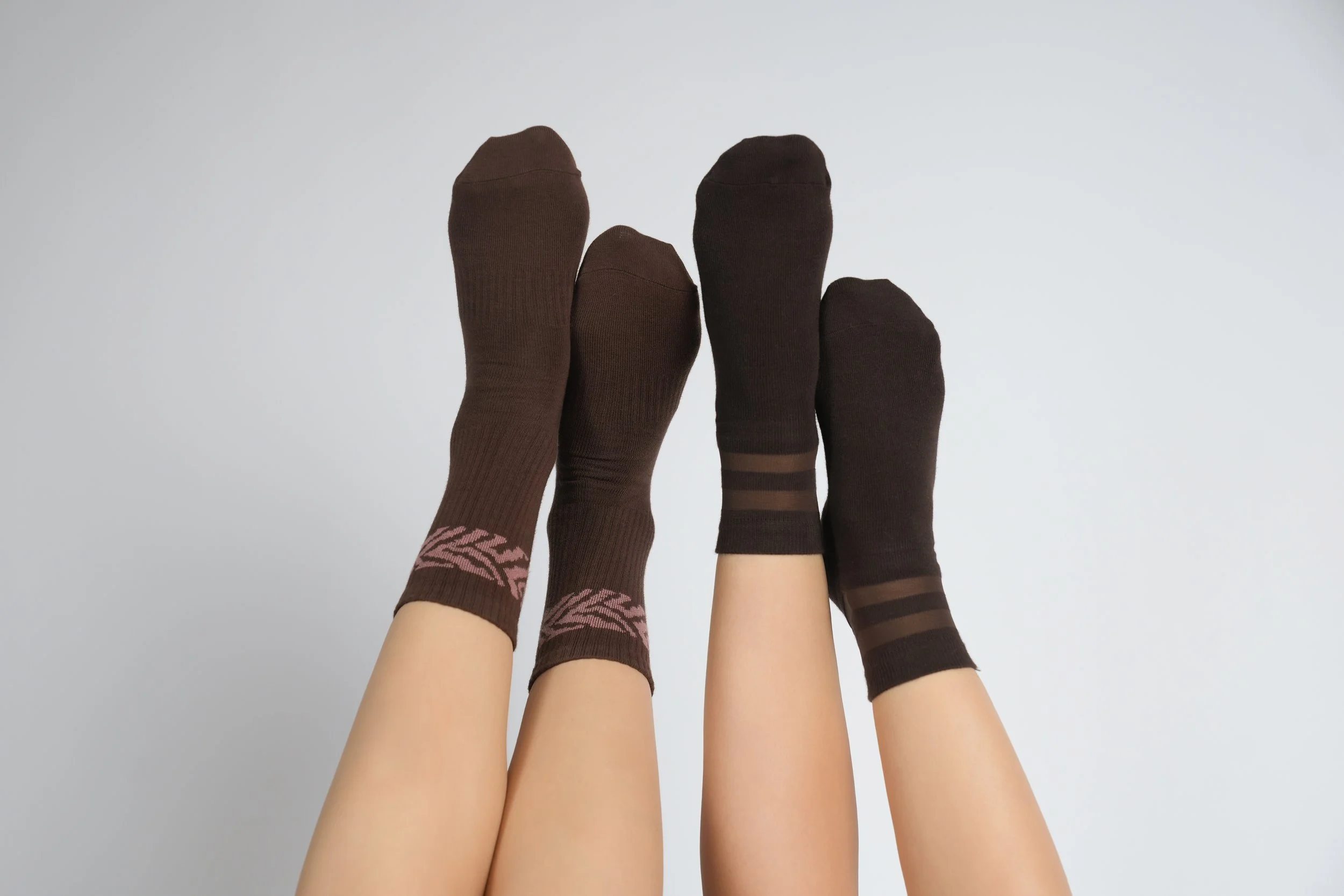 Four feet with legs raised wearing two different pairs of brown grip socks against a plain white background.