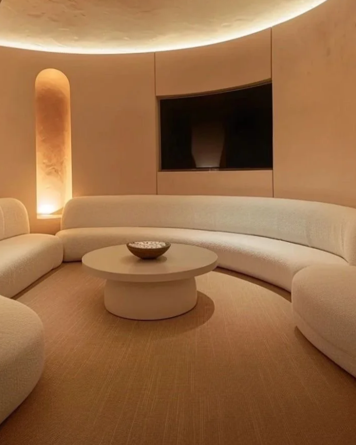 The present healing space experience hub, first in the world, Minimalist living room with curved beige sofa, round coffee table, and a casting lighted niche in the wall.