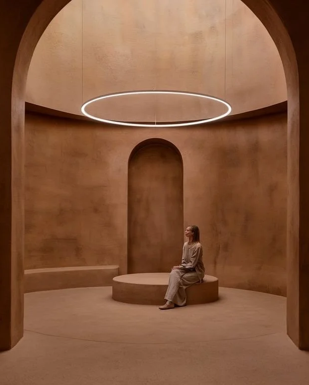 The present healing space experience hub, unity light, A woman sitting on a circular bench in a minimalist, modern interior with earthy tones and a large circular light fixture hanging from the ceiling.