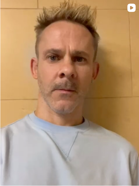 Dominic Monaghan - A man with short, spiked blonde hair, wearing a light blue shirt, looking directly at the camera with a serious expression, standing in front of a light-colored wooden wall.