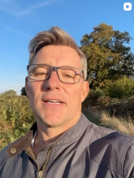 Ben Shepherd - A man wearing glasses and a gray jacket outdoors on a sunny day, with trees and a blue sky in the background.