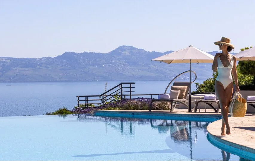 A woman in a white swimsuit, straw hat, and sunglasses walking by a swimming pool, holding a straw bag and a towel, with lounge chairs and umbrellas, overlooking a lake and mountains in the background on a sunny day.